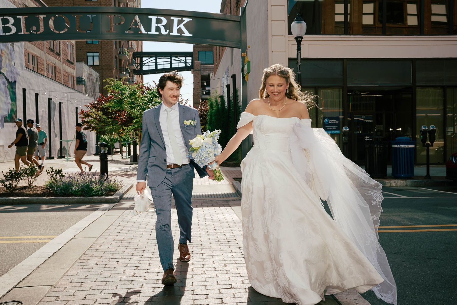 Bride and groom hold hands running across city street near Bijou Park on wedding day.
