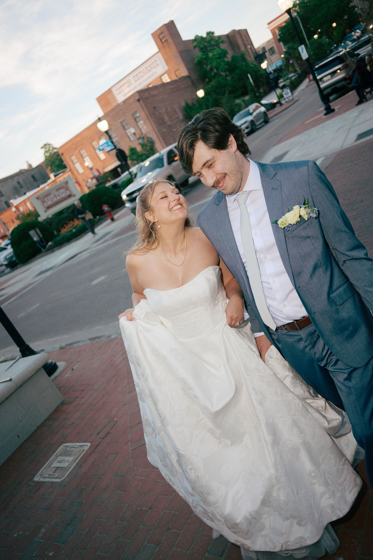 A smiling bride in a strapless white gown and groom in a gray suit walk together on a brick sidewalk in a city.