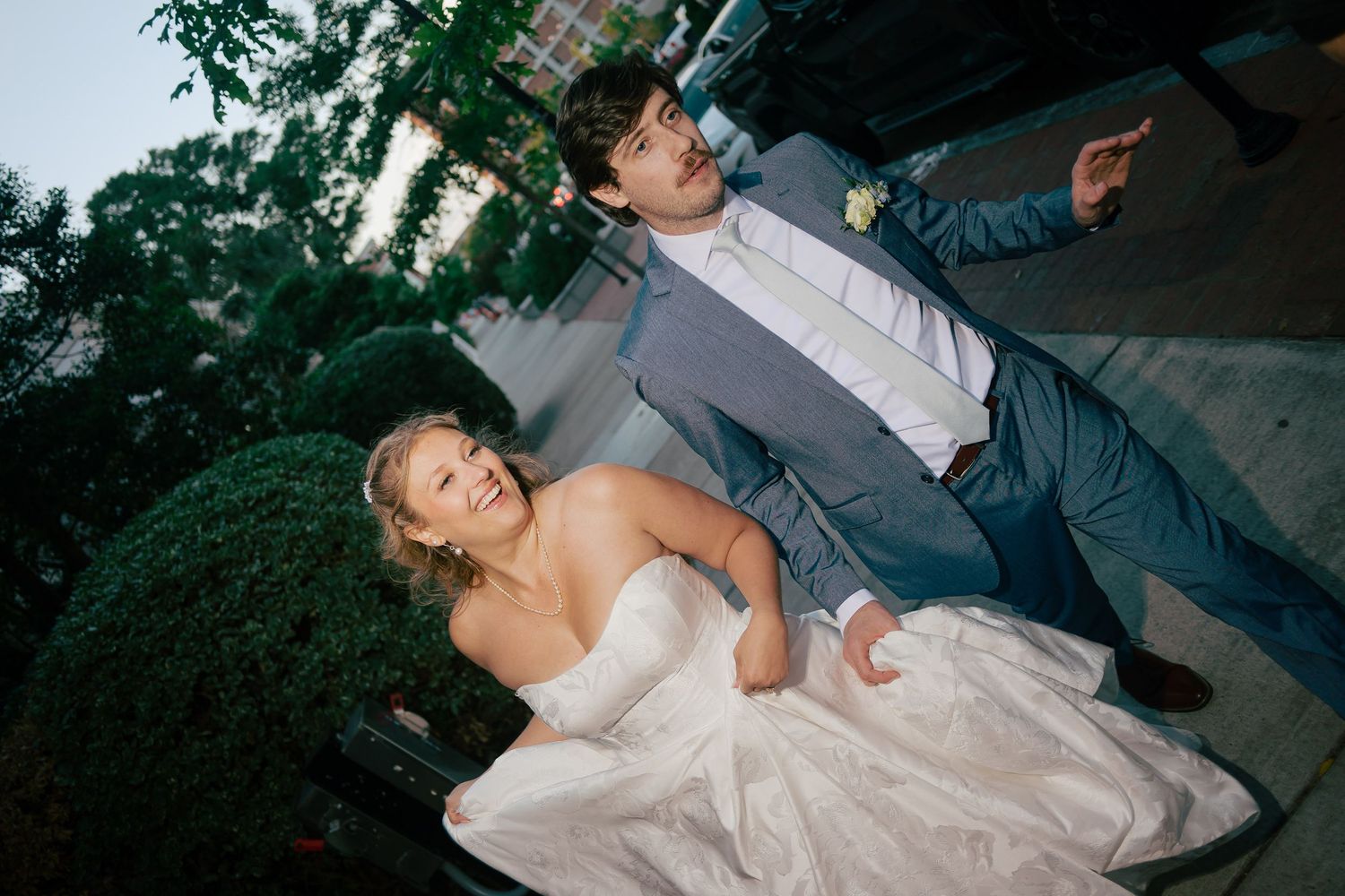 Bride and groom posing playfully outdoors at dusk near lush green hedges in wedding attire.