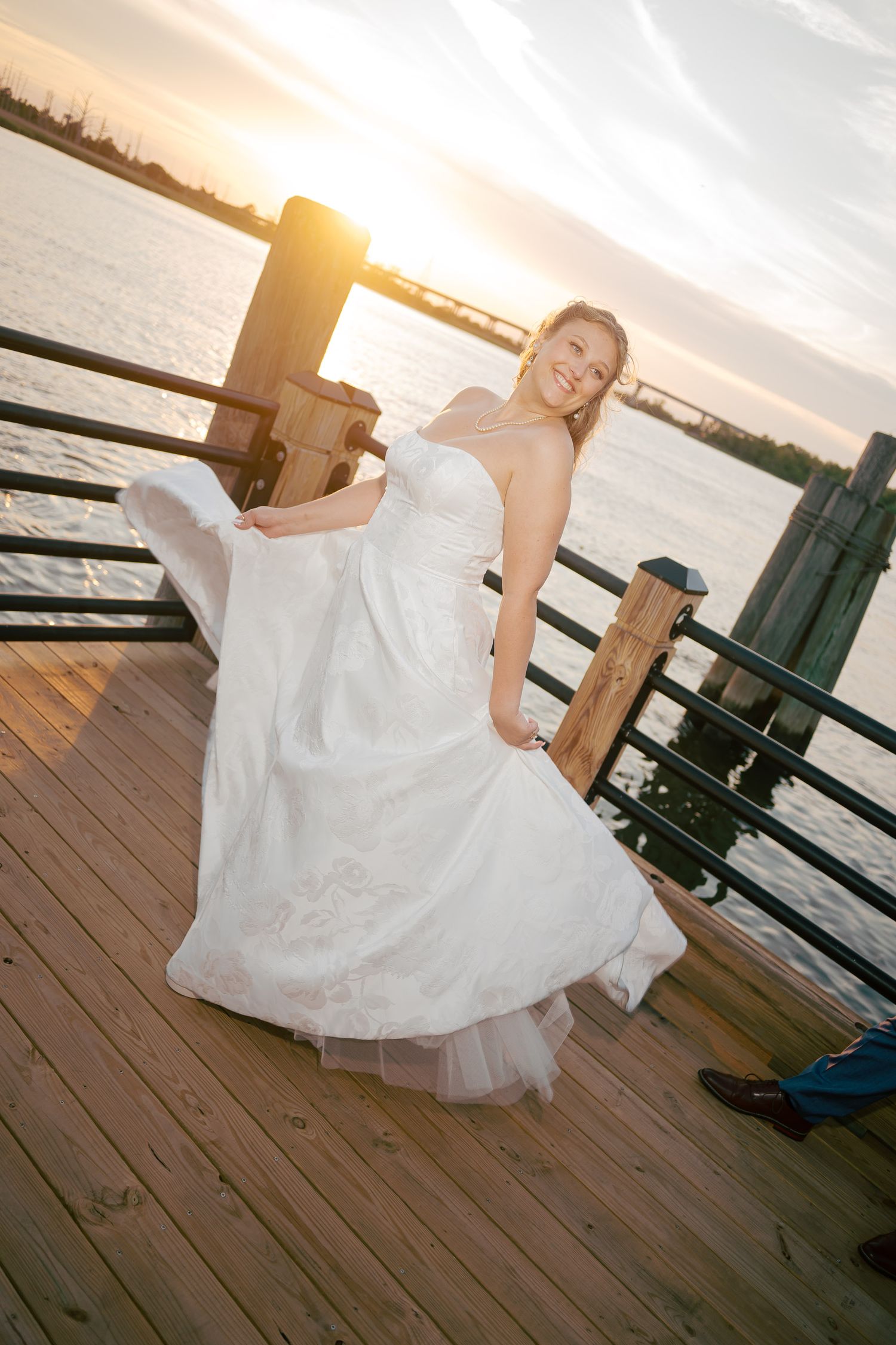 Bride in strapless white gown twirls on wooden pier at golden sunset over waterway.