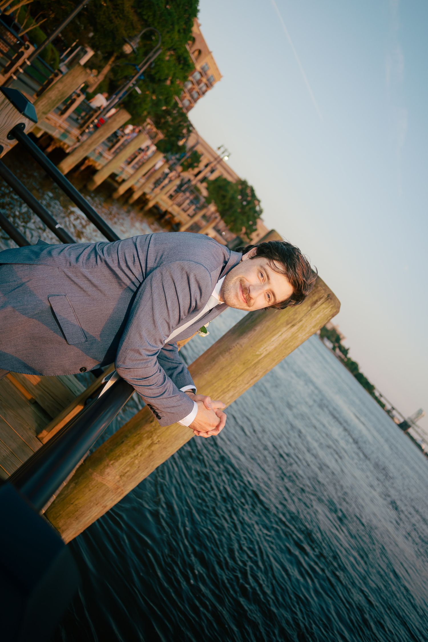 Dapper groom in a gray suit leaning on a wooden dock railing with a waterfront view at sunset.