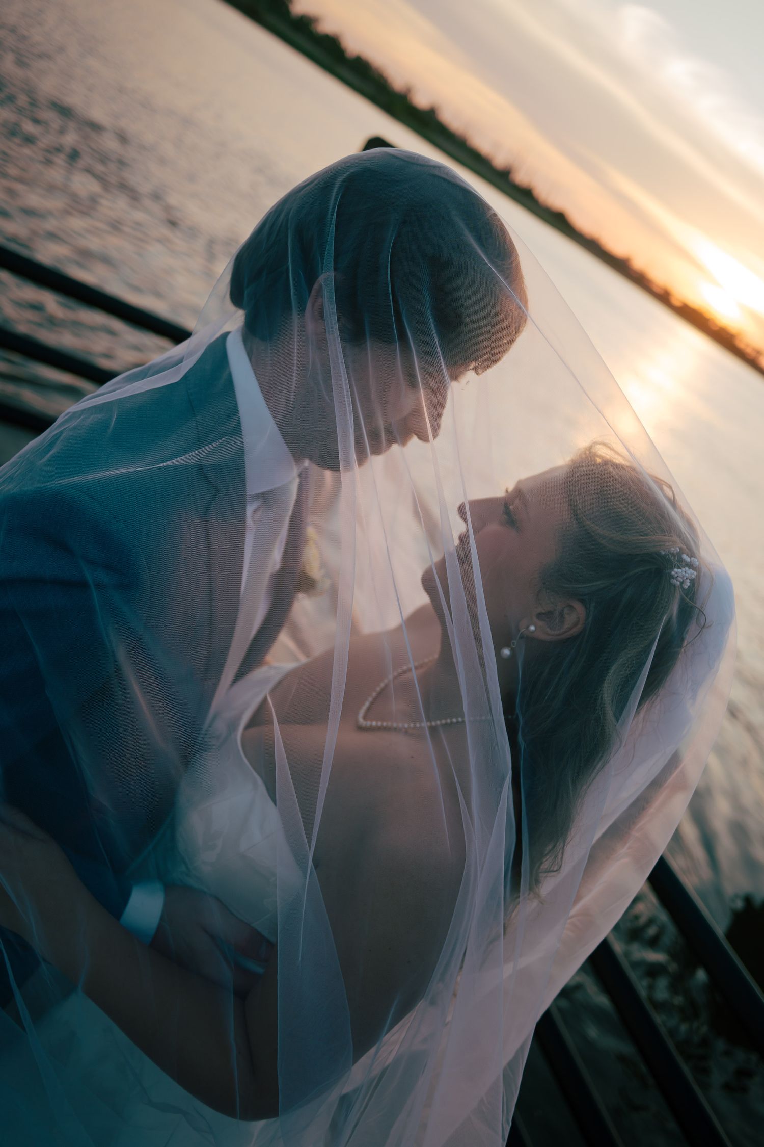 Bride and groom sharing romantic moment under veil at sunset by waterfront on wedding day.
