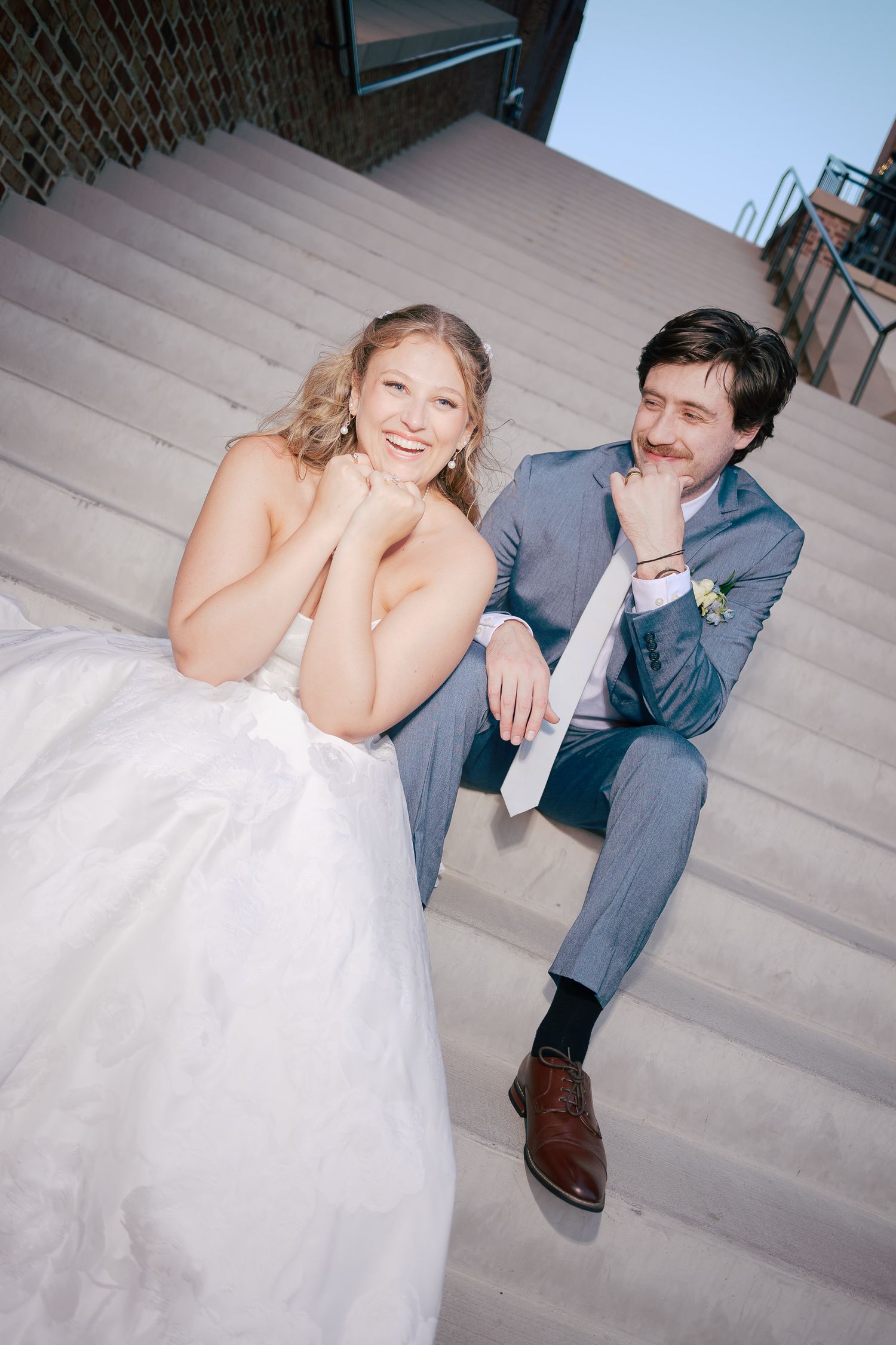 Bride and groom in gray suit sitting together on outdoor steps, smiling during wedding portraits.