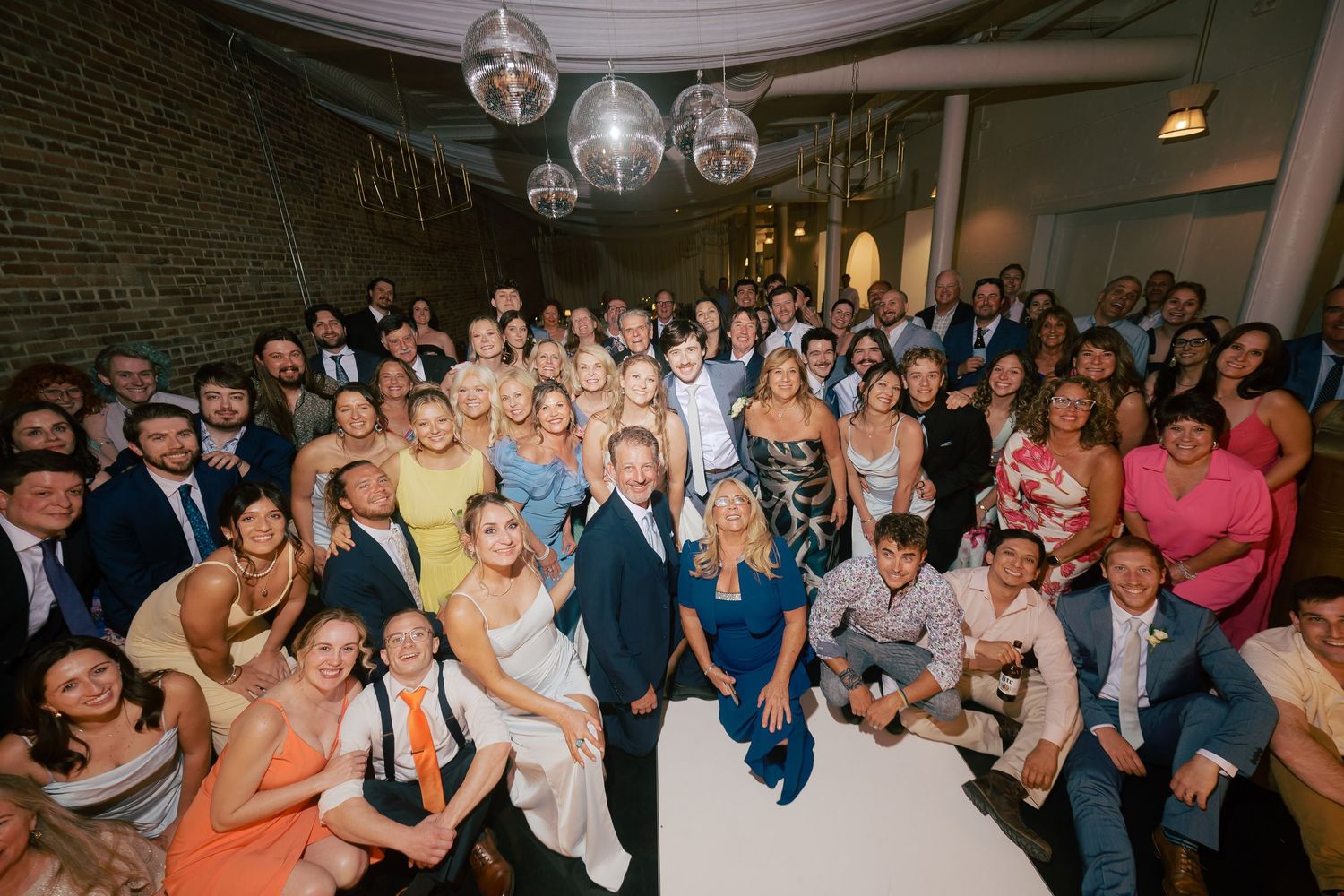 Large wedding reception group photo with smiling guests gathered together in a decorated venue with pendant lights.