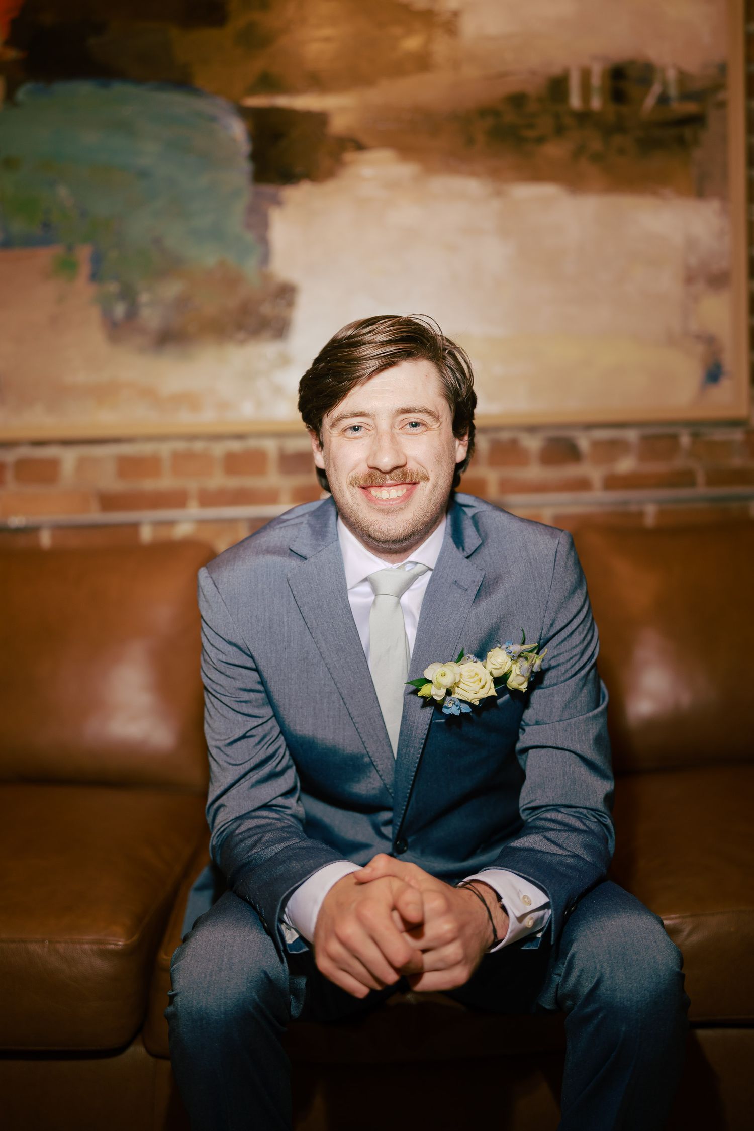 Smiling groom in gray suit with boutonniere sits on leather sofa at wedding venue.