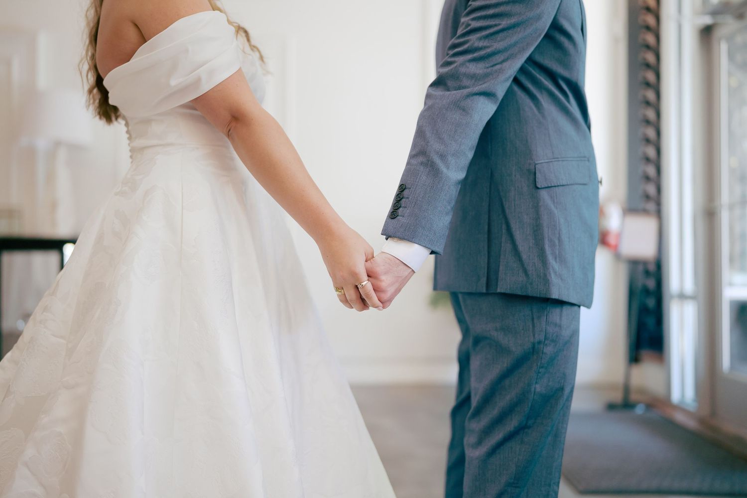 Bride in white off-shoulder gown and groom in gray suit holding hands at wedding ceremony.