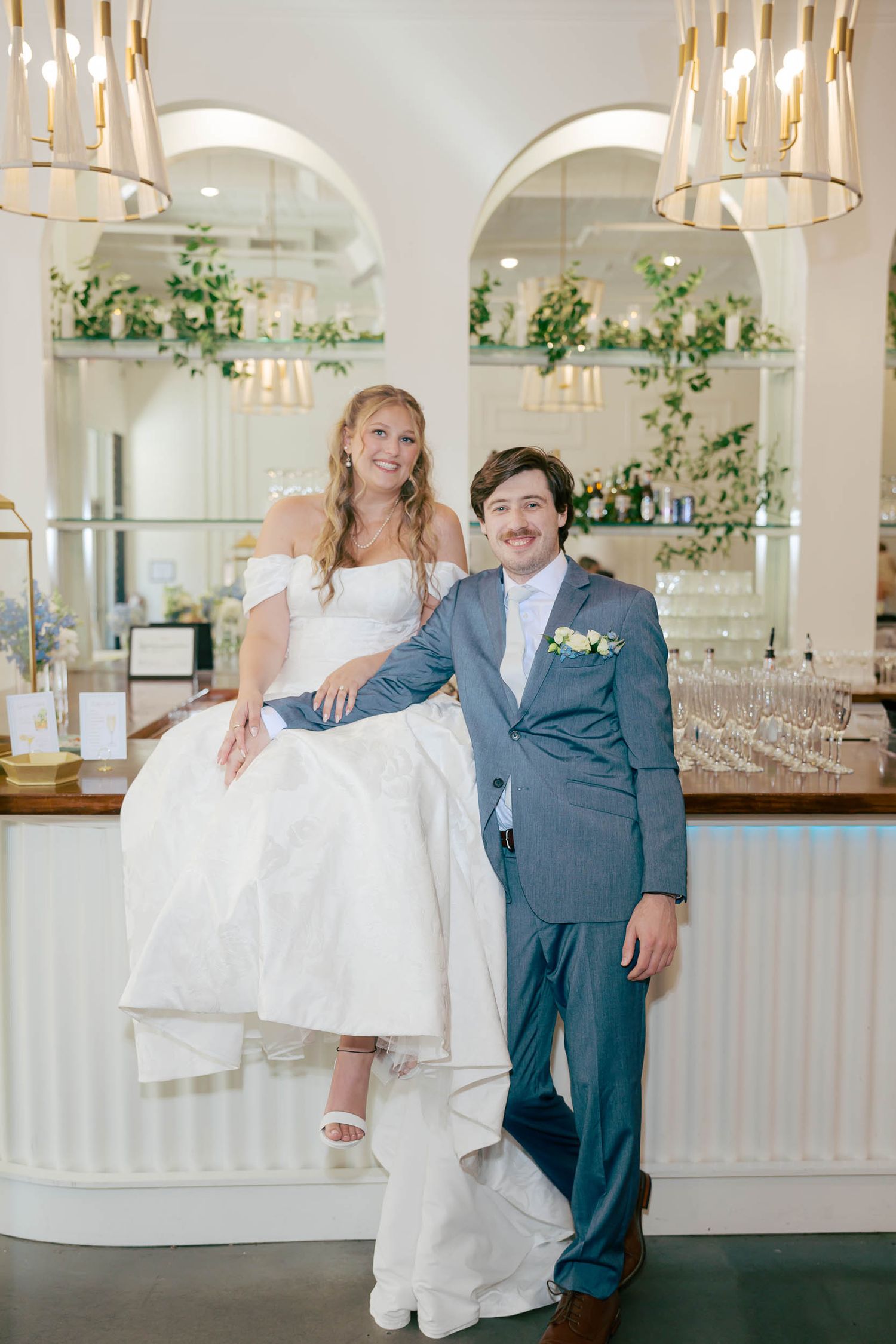 Bride and groom posing together at elegant wedding venue bar, bride seated in white gown, groom in navy suit.