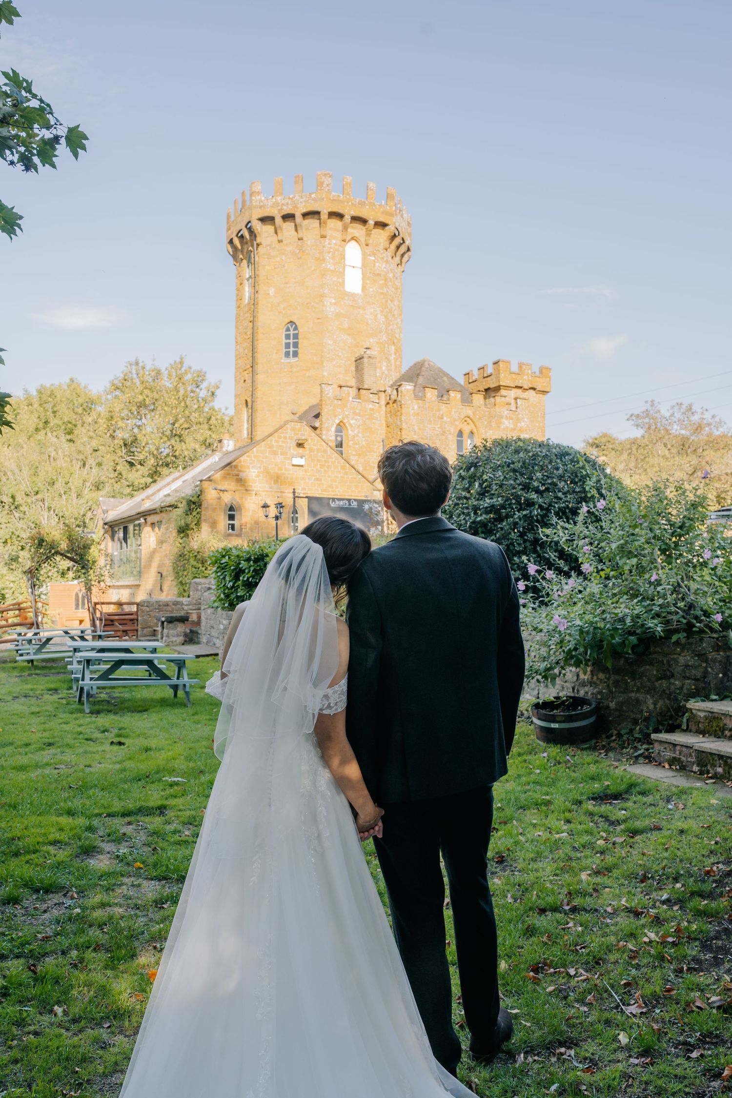 Bride and groom holding hands facing a historic stone castle tower on their wedding day.