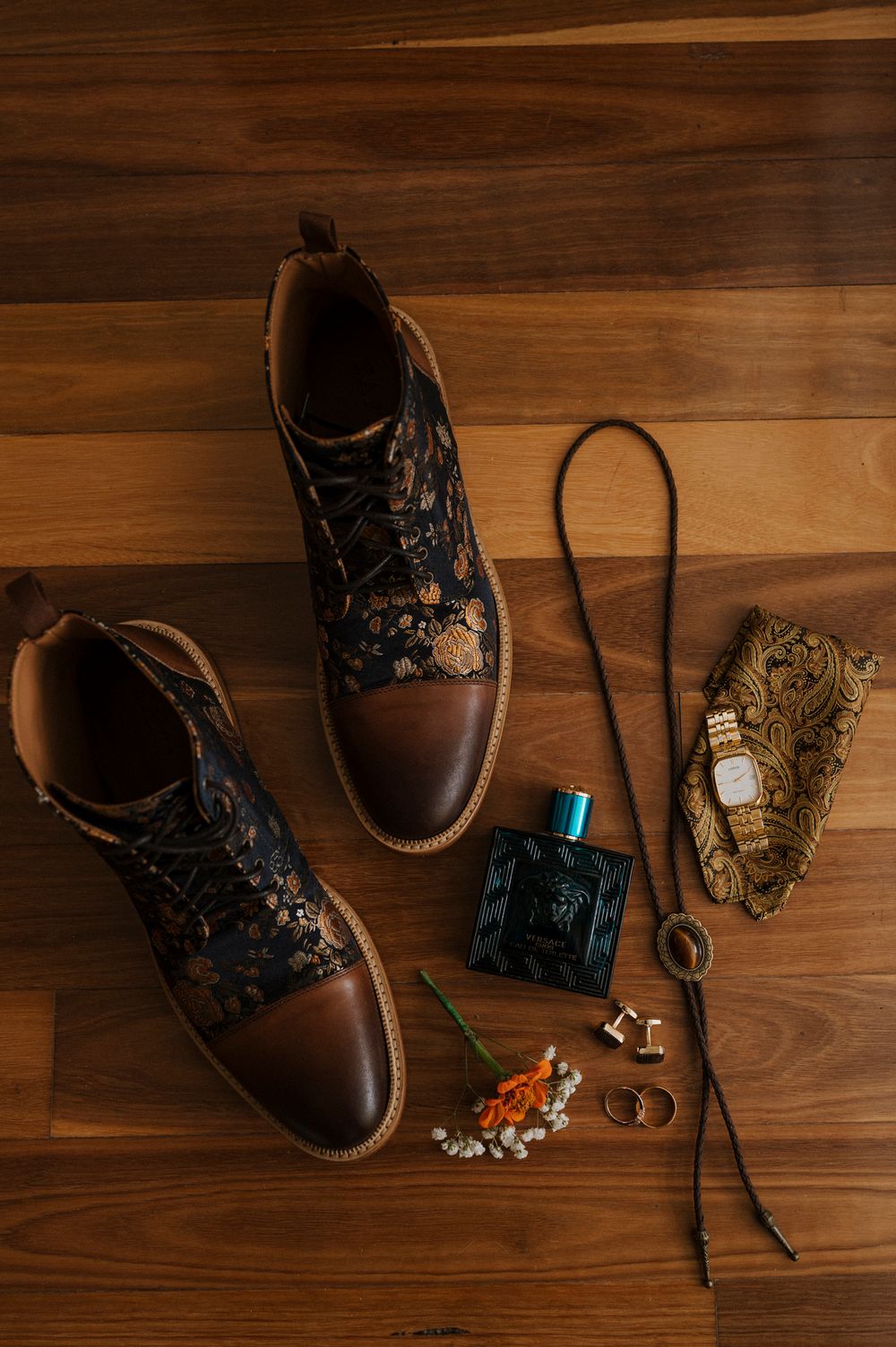 Brown leather floral boots with cologne, cufflinks, bolo tie, rings, and pocket square on wood floor.