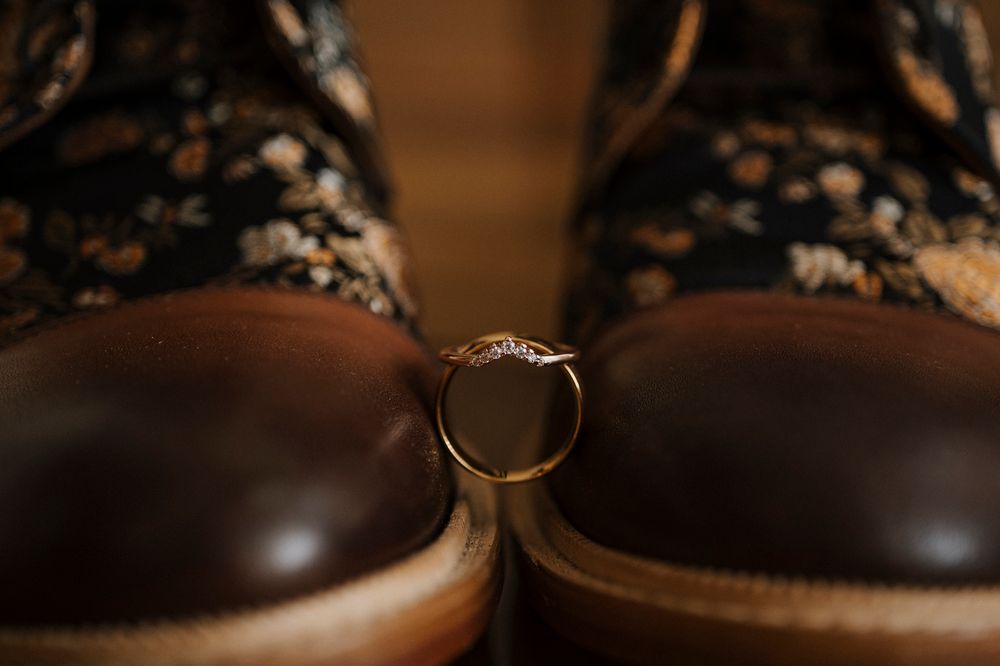 A gold wedding ring resting between two brown leather dress shoes with floral fabric detailing in warm ambient light.