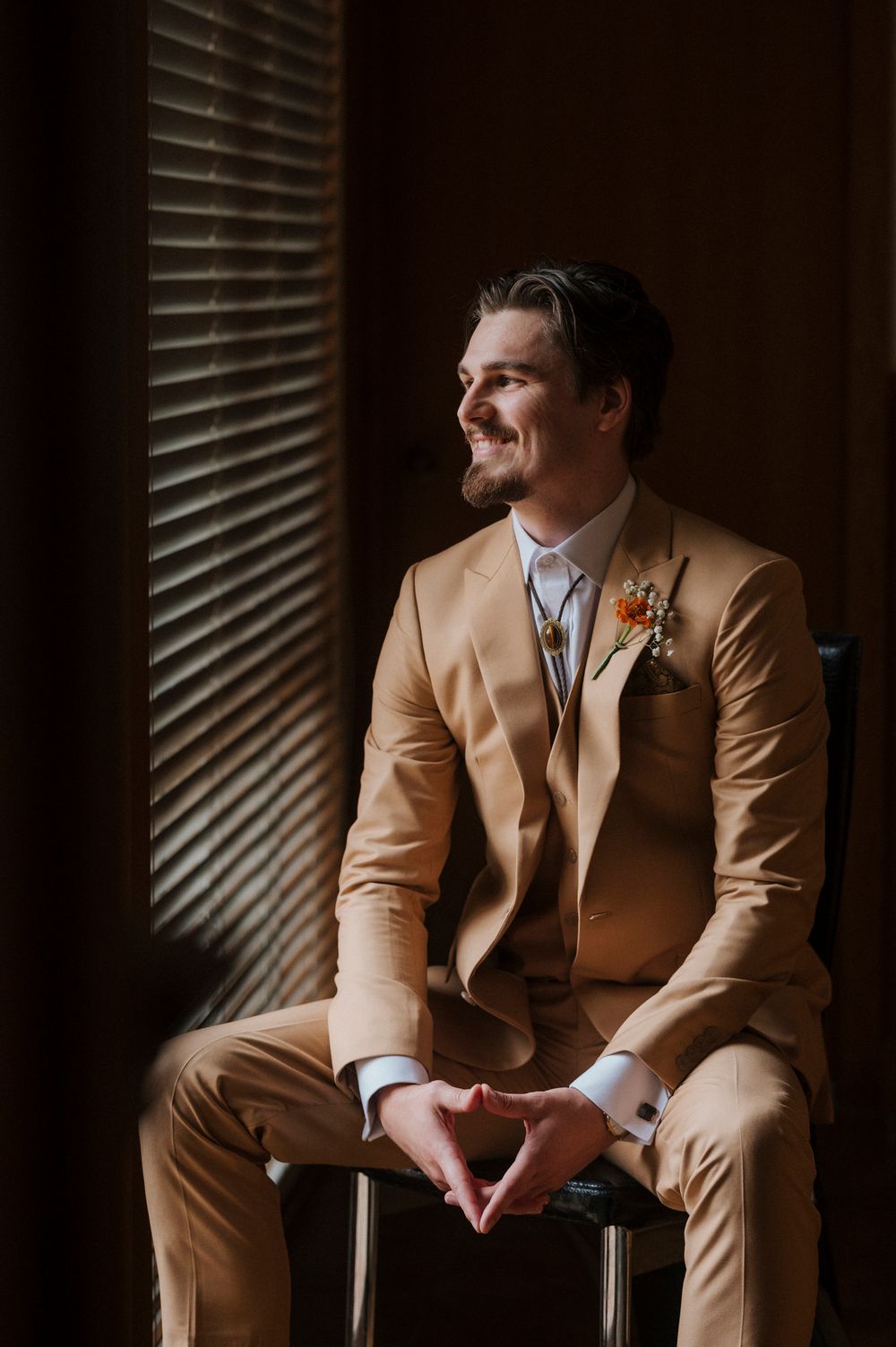 A groom in a tan suit with a floral boutonniere sits smiling by window blinds on his wedding day.