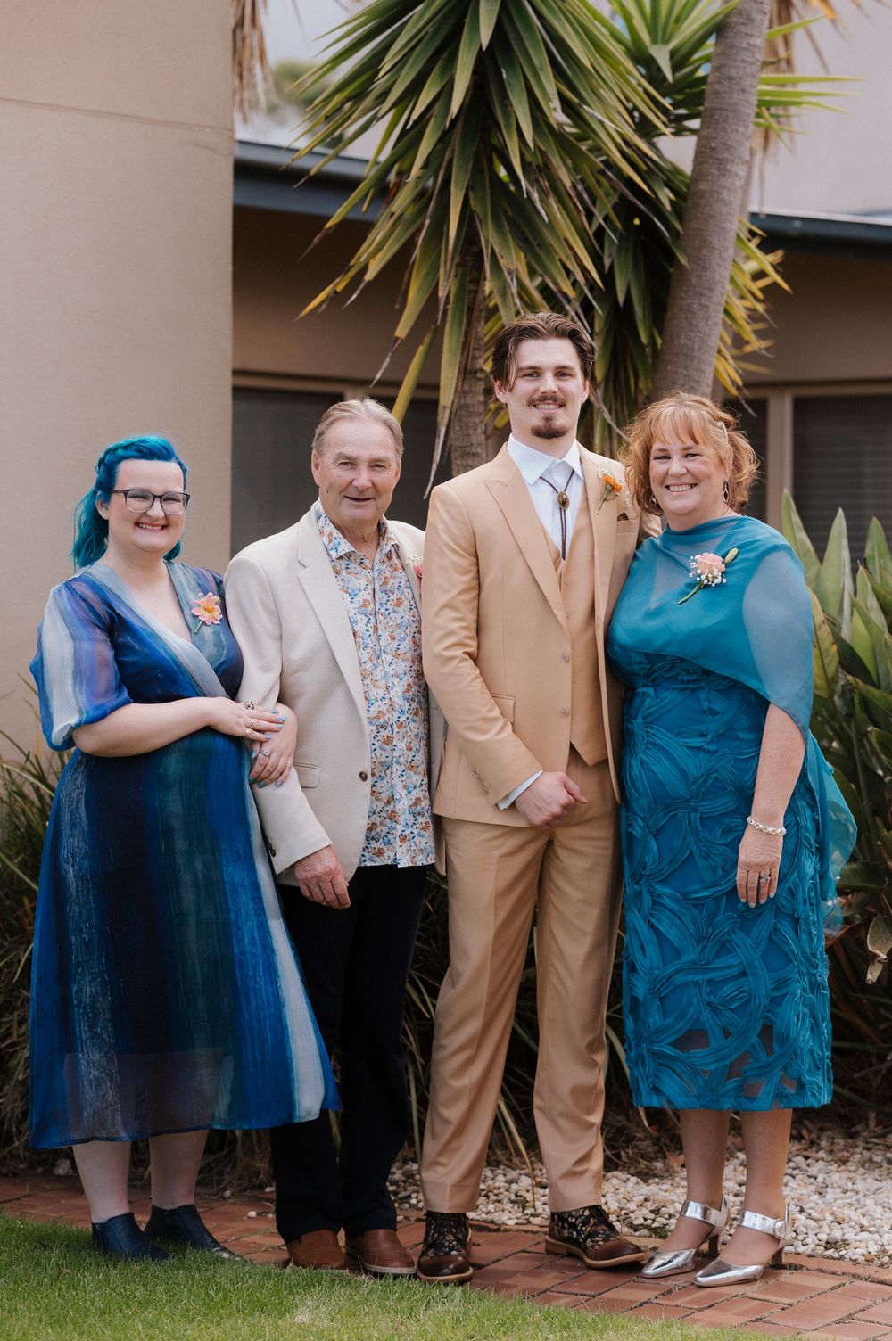 Family of four posing outdoors at a wedding, featuring a groom in tan suit with guests in blue and teal formal attire.