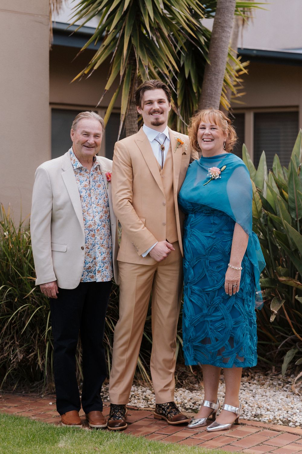 A groom in a tan suit poses with his parents, father in floral shirt and mother in teal dress, outdoors near palm trees.