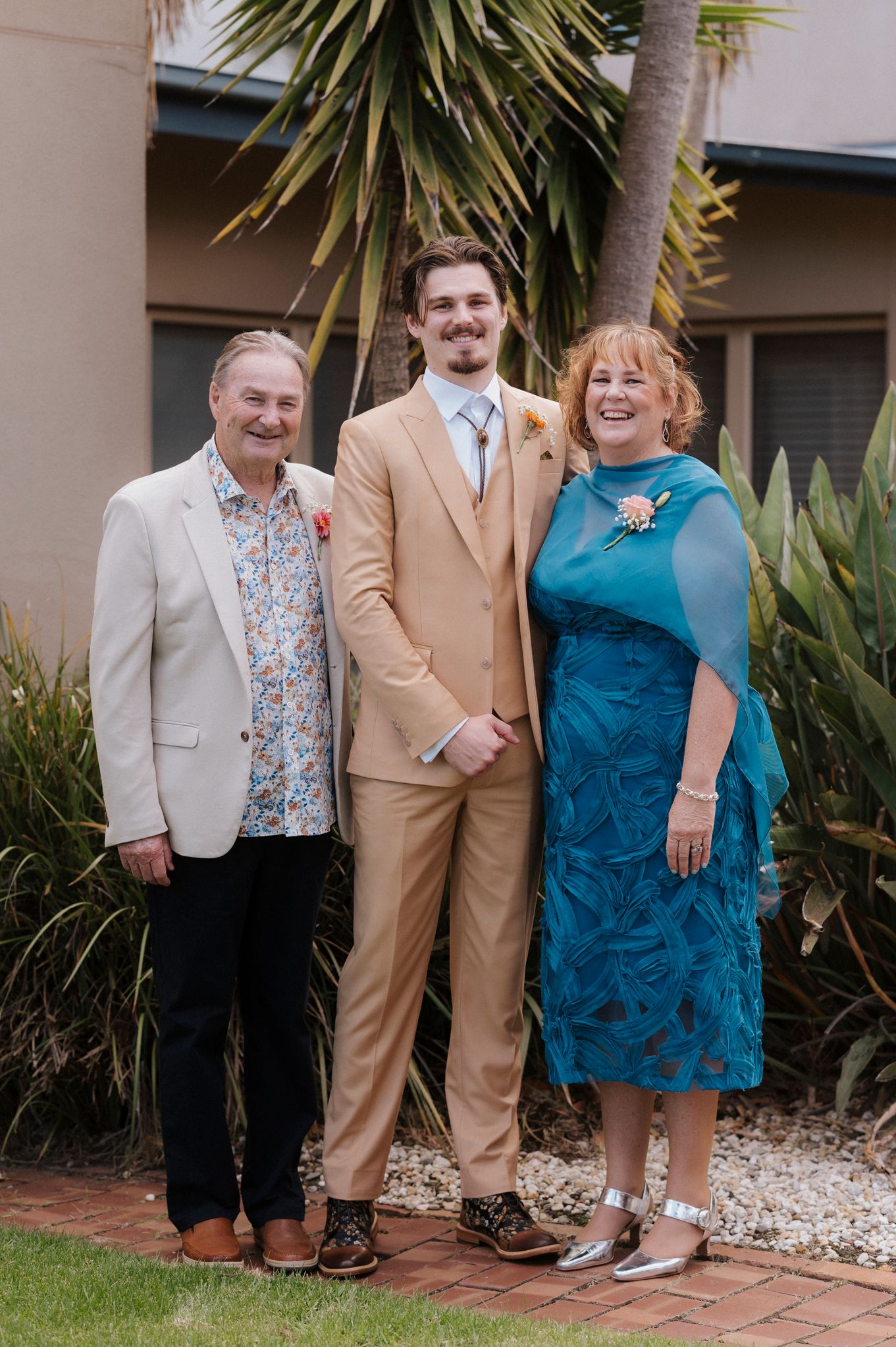 A groom in a tan suit poses with his parents, father in floral shirt and mother in teal dress, outdoors near palm trees.