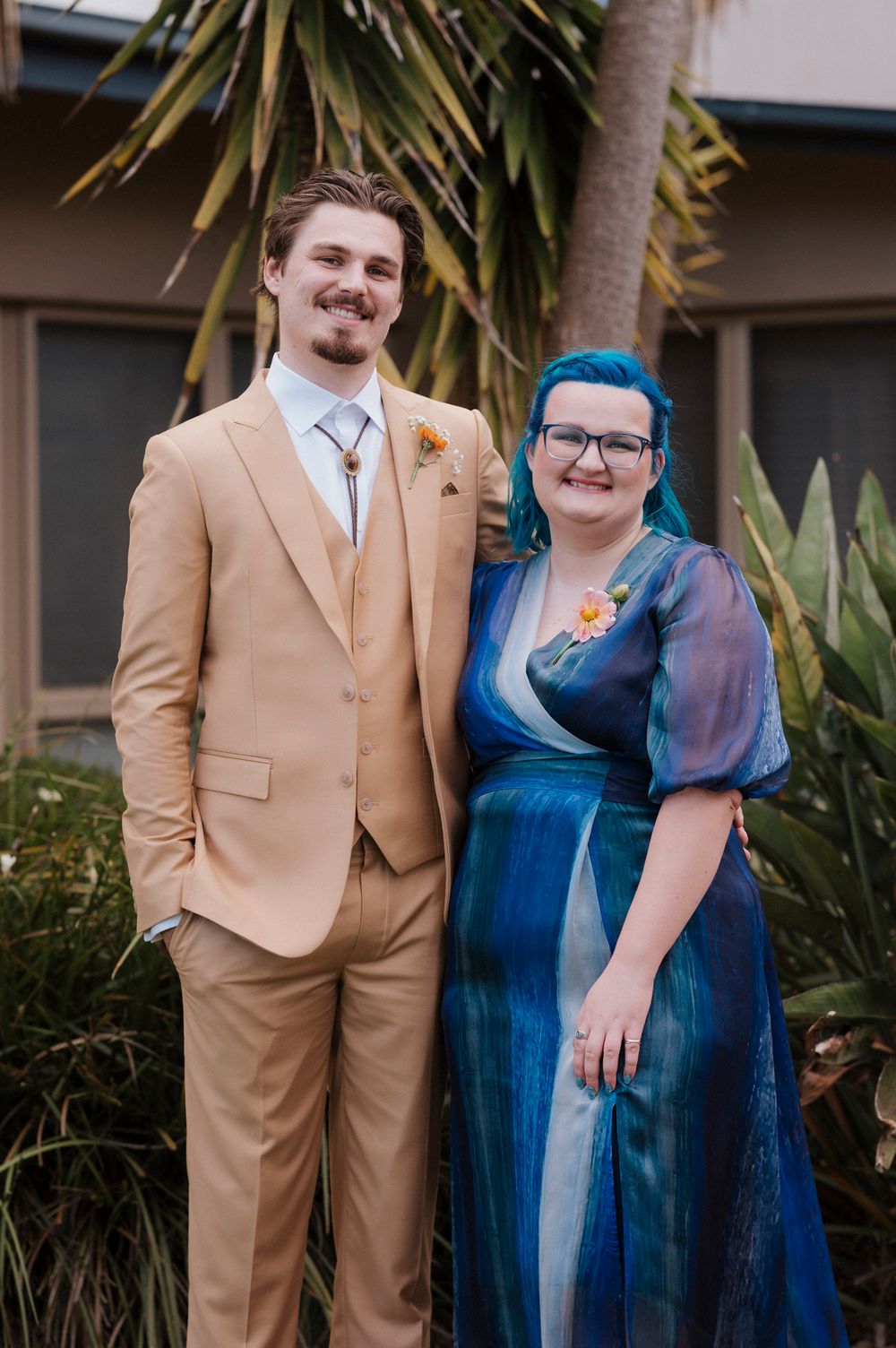 A man in a tan suit and woman in a blue dress with teal hair pose together at a formal event outdoors.