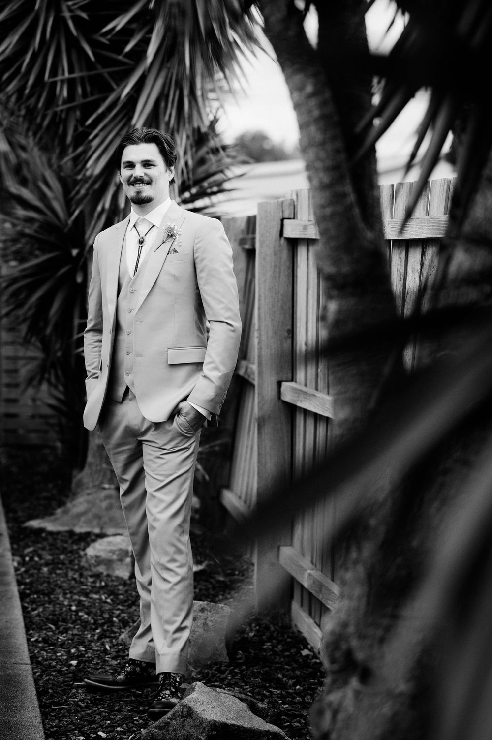 Black and white photo of a groom in a light suit leaning against a wooden fence surrounded by tropical palms.