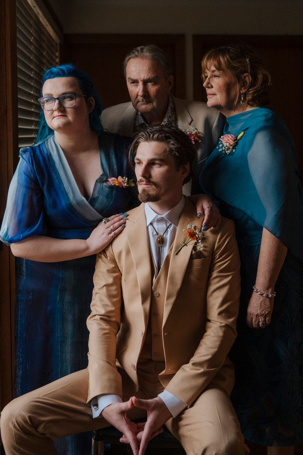 Groom in tan suit seated with family in blue attire posing for a formal wedding portrait indoors.
