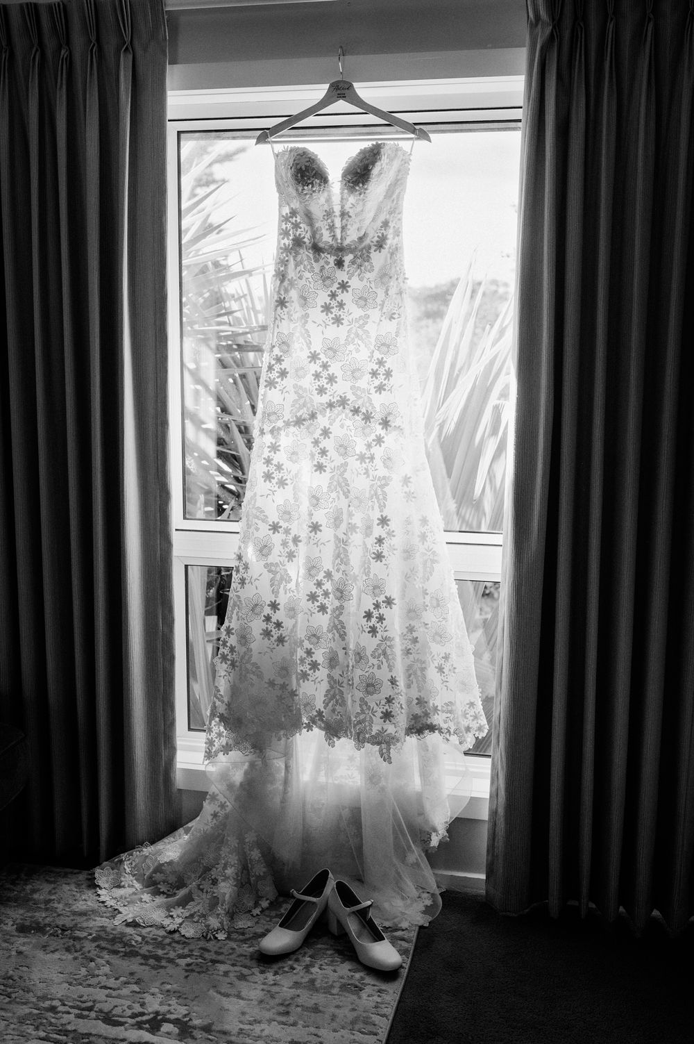 Black and white photo of an ornate strapless wedding gown with floral lace details hanging in a window with bridal shoes below.