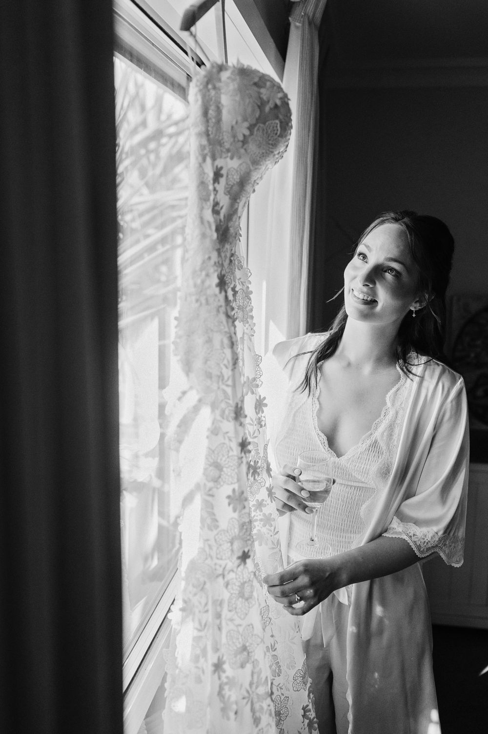 Bride in white lace robe holding champagne glass by window with wedding dress hanging nearby, black and white photo.