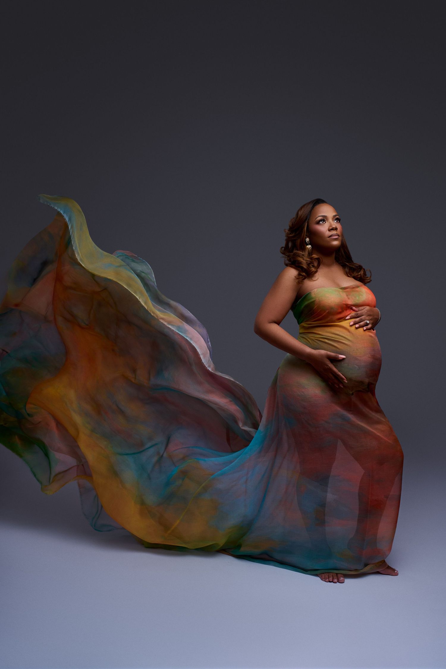 Pregnant woman in flowing multicolored fabric gown posing gracefully against gray studio background.