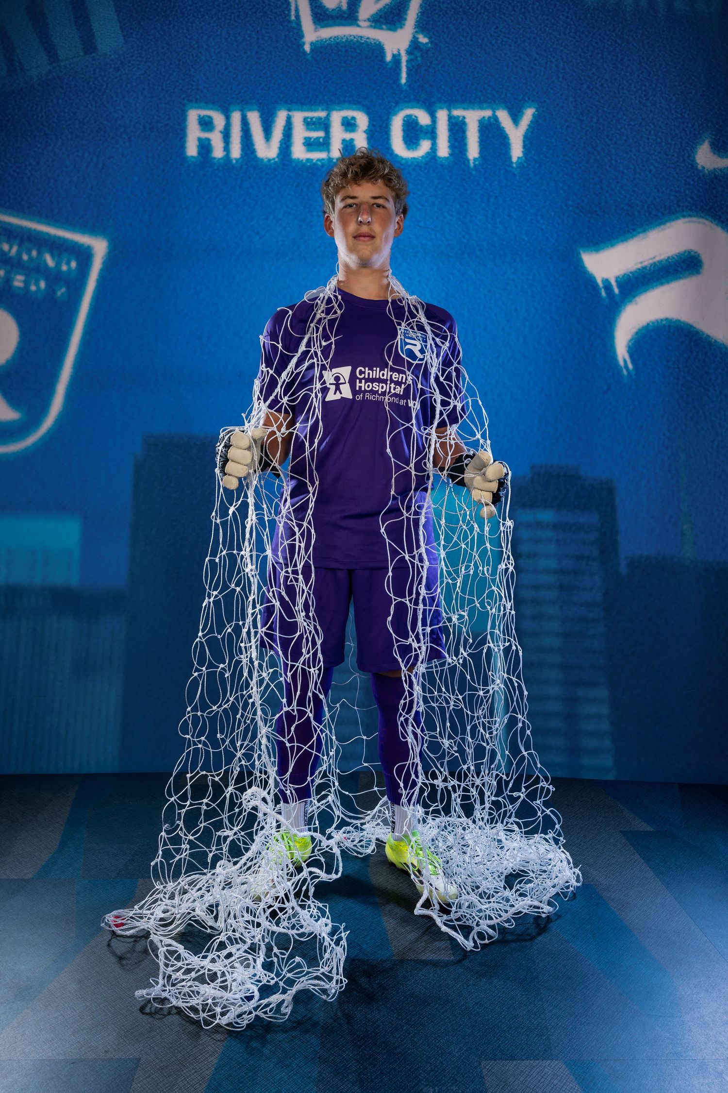 A young man in a purple Children's Hospital shirt holds a soccer goal net at a River City FC themed backdrop.
