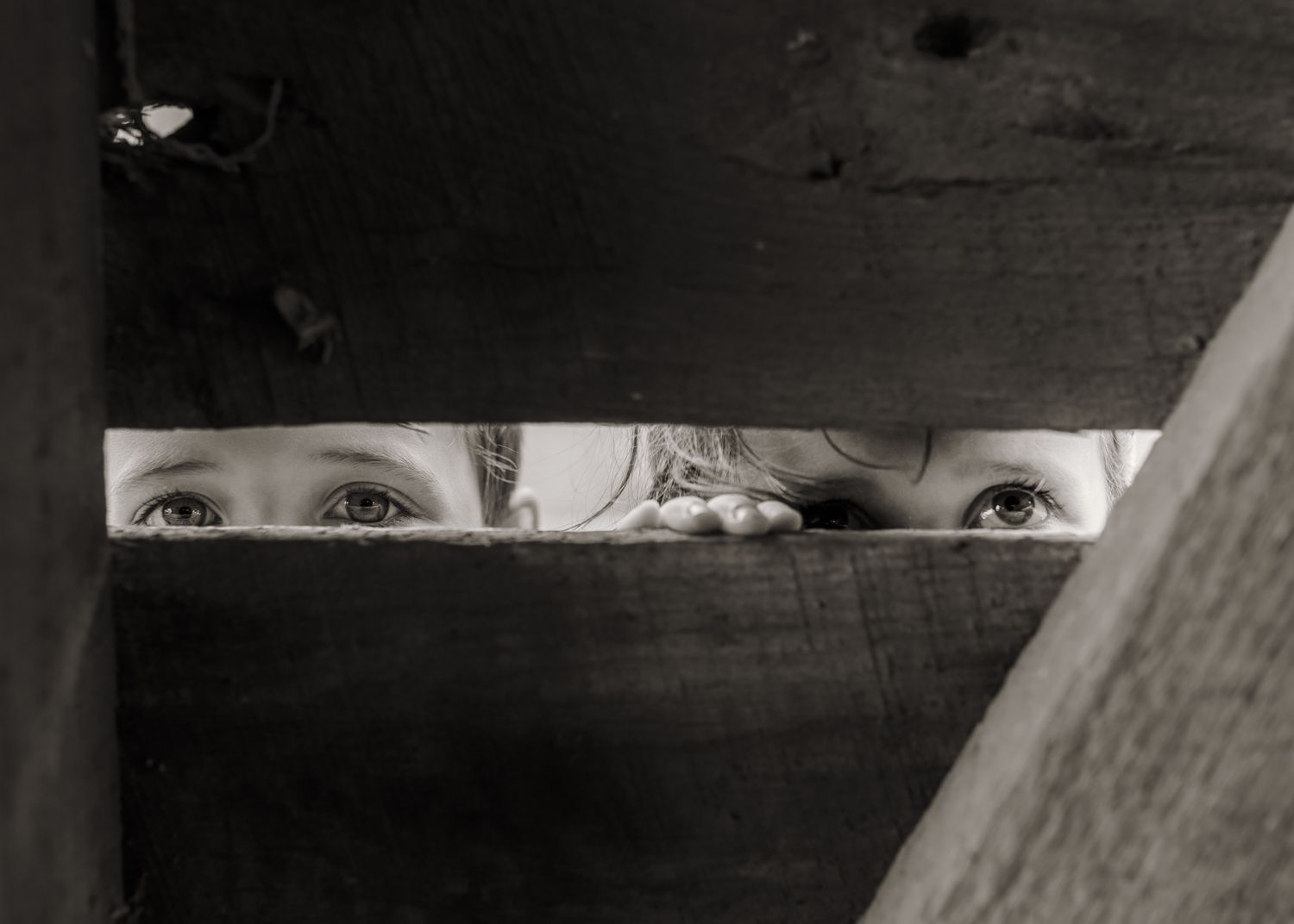 Black and white photo of a child's eyes peeking through a narrow gap between wooden boards.