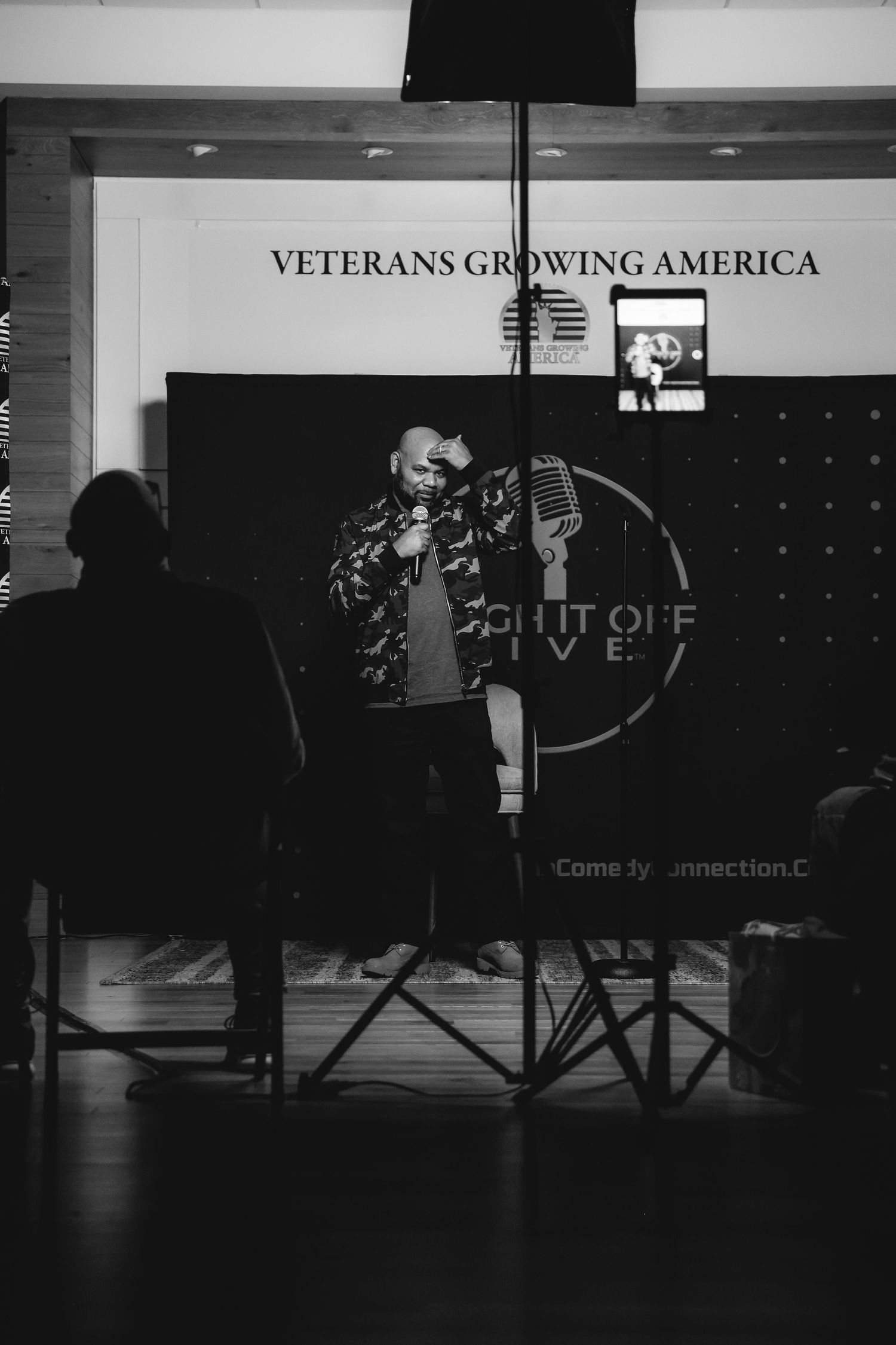 Bald man in camo jacket performs stand-up comedy at microphone stand on wooden stage floor.