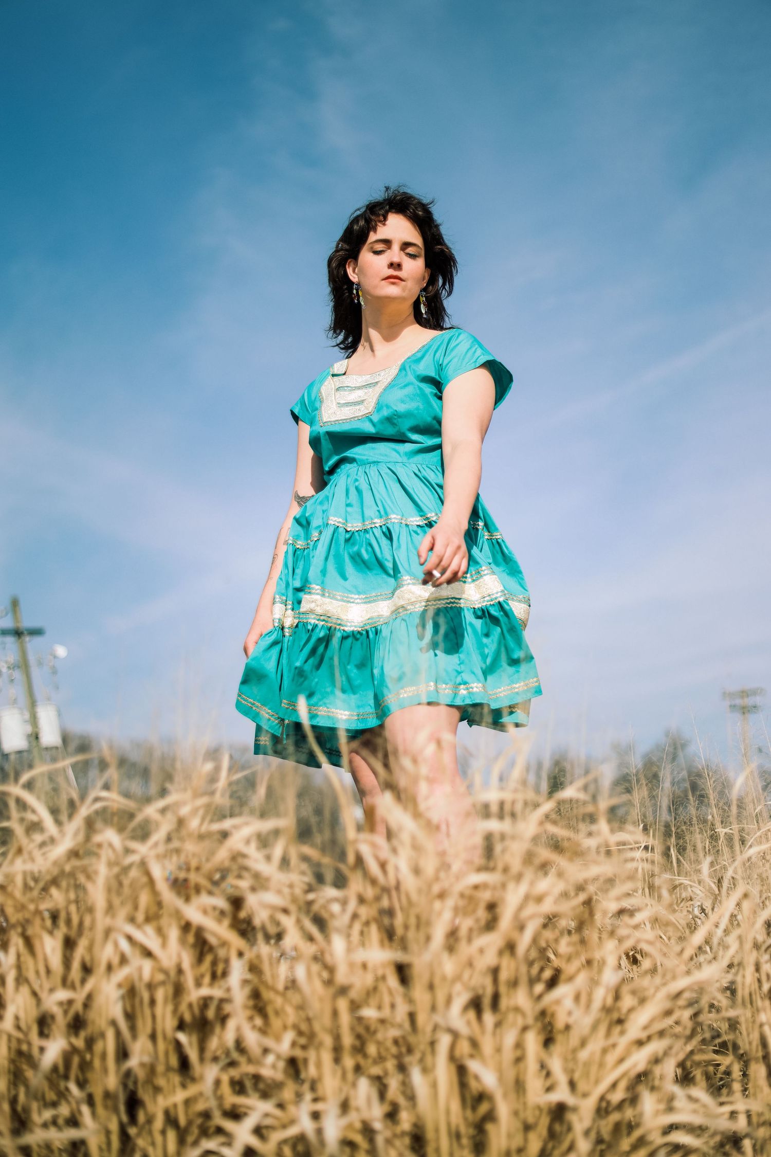 Woman in teal vintage dress standing in golden wheat field under blue sky.