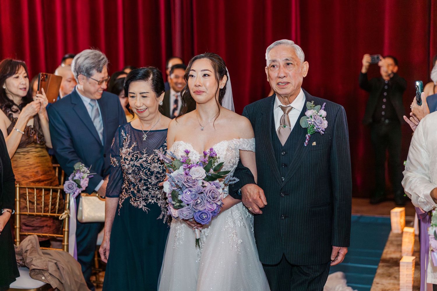 Bride in off-shoulder gown holding purple bouquet walks down aisle with elderly man and woman at wedding ceremony.