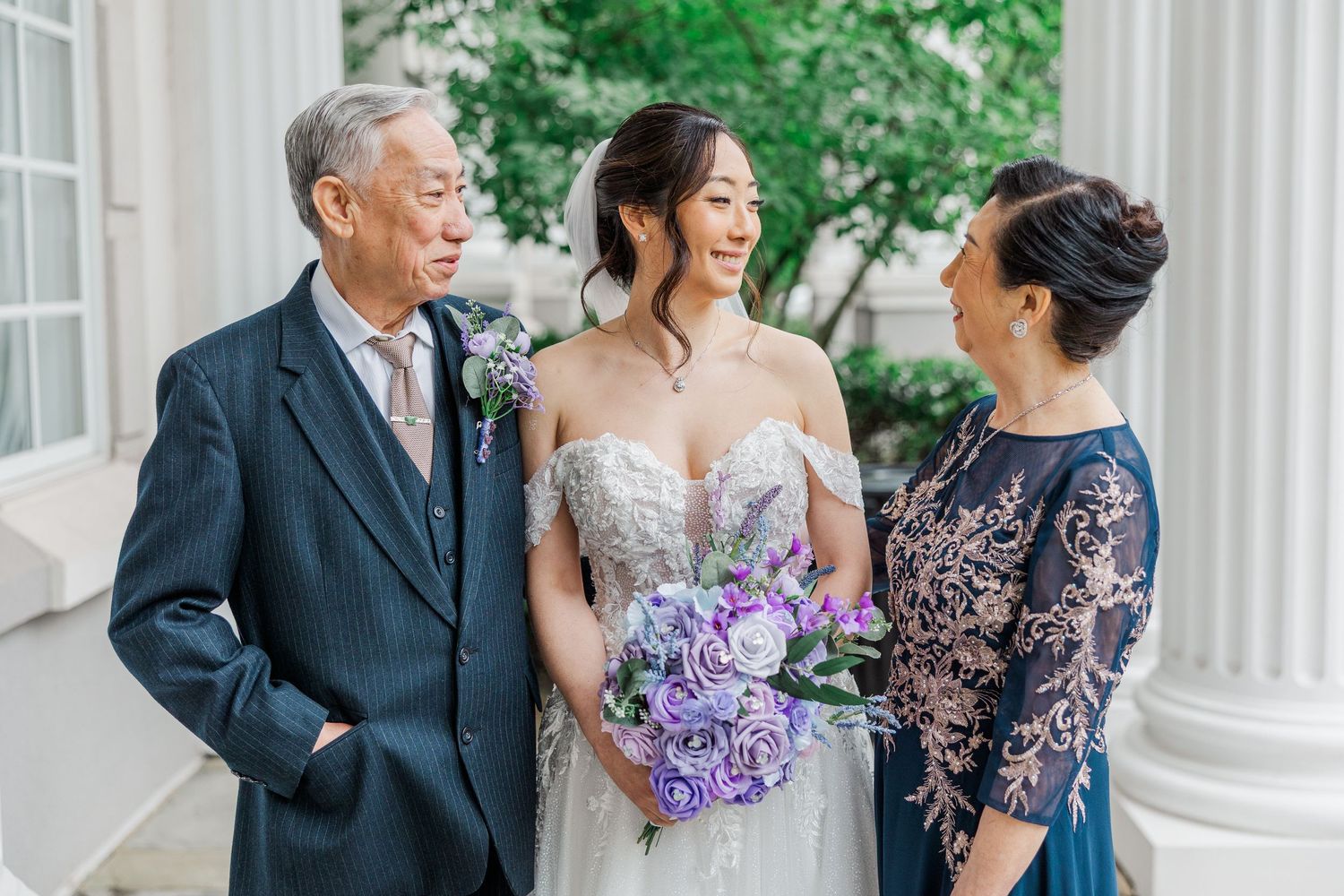 Bride in lace gown holds purple bouquet, smiling with elderly parents at elegant white column venue.