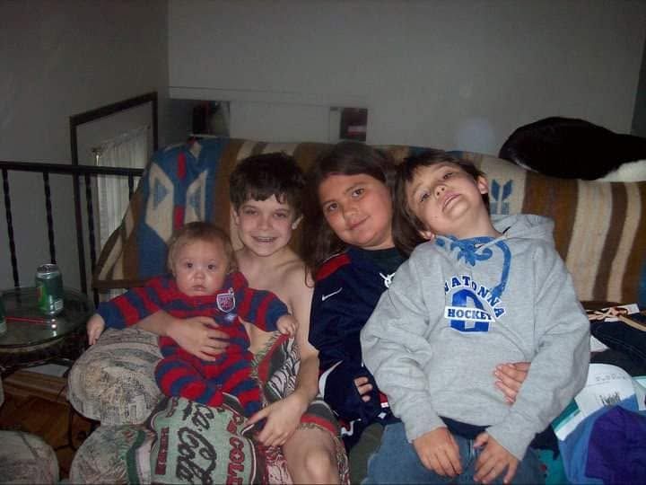 Four young children sitting together on a couch, smiling and posing for a casual family photo indoors.