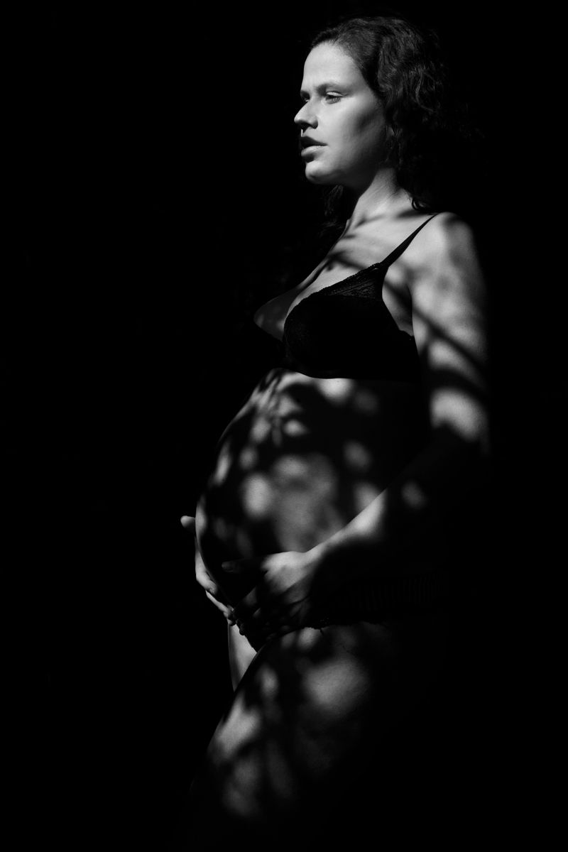Black and white portrait of a pregnant woman in lingerie with dramatic leaf shadows on her body.