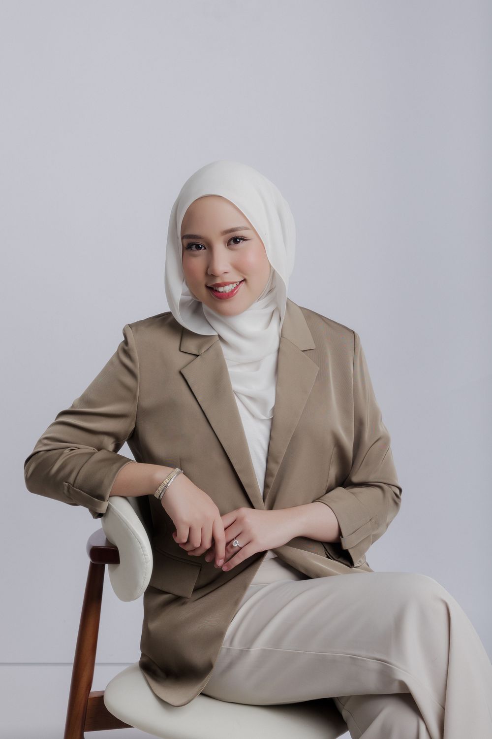 A smiling woman in a white hijab and beige blazer sits elegantly on a chair against a light background.