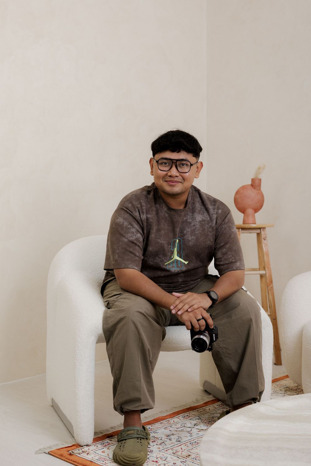 Young man in Jordan t-shirt and glasses sits in white armchair holding camera in minimalist room.