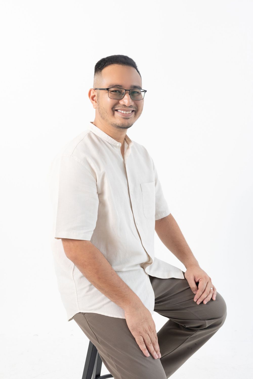 Smiling Asian man in glasses and white shirt sitting on stool against white background.