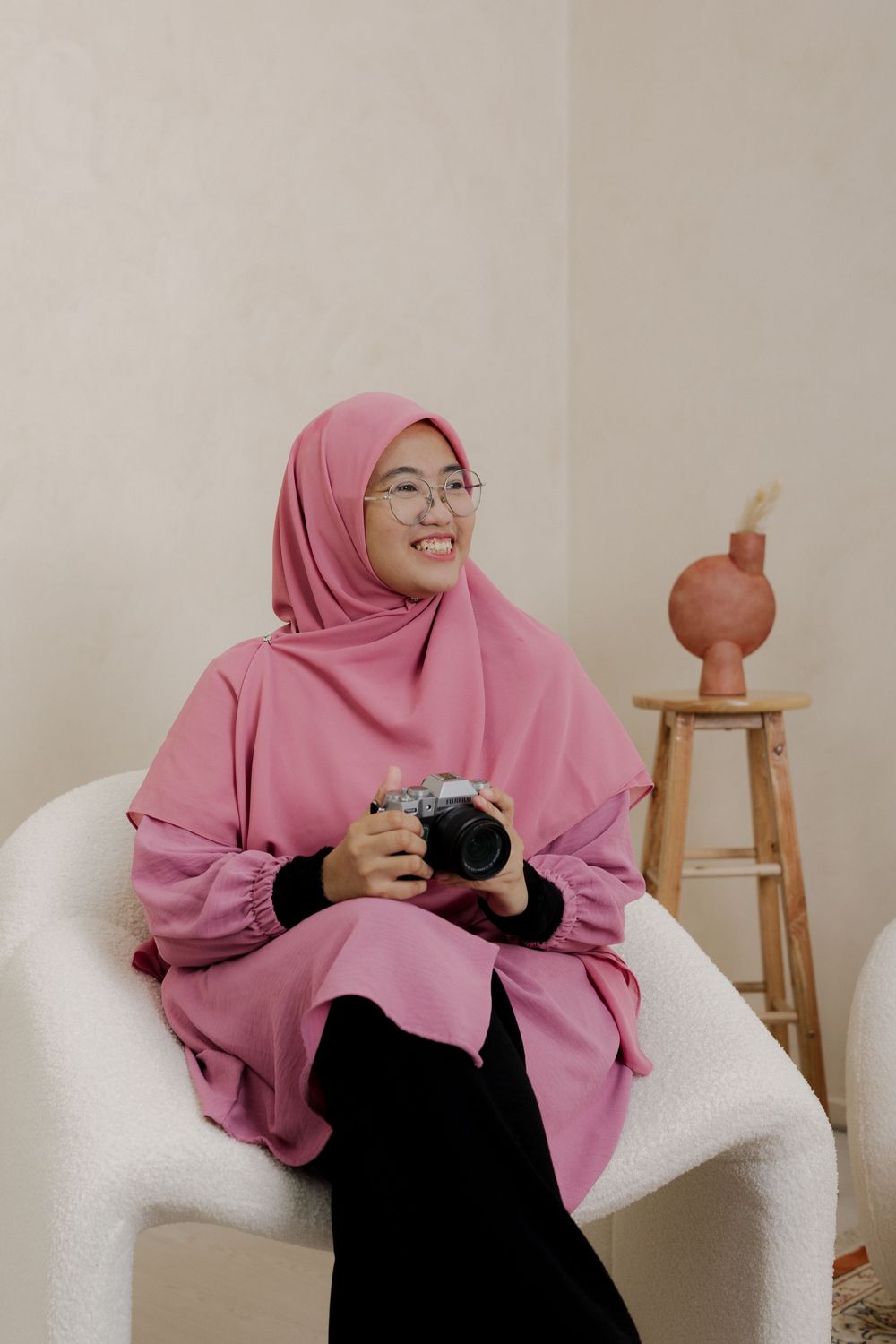 Smiling woman in pink hijab and glasses sitting in white chair holding a camera in a bright studio setting.