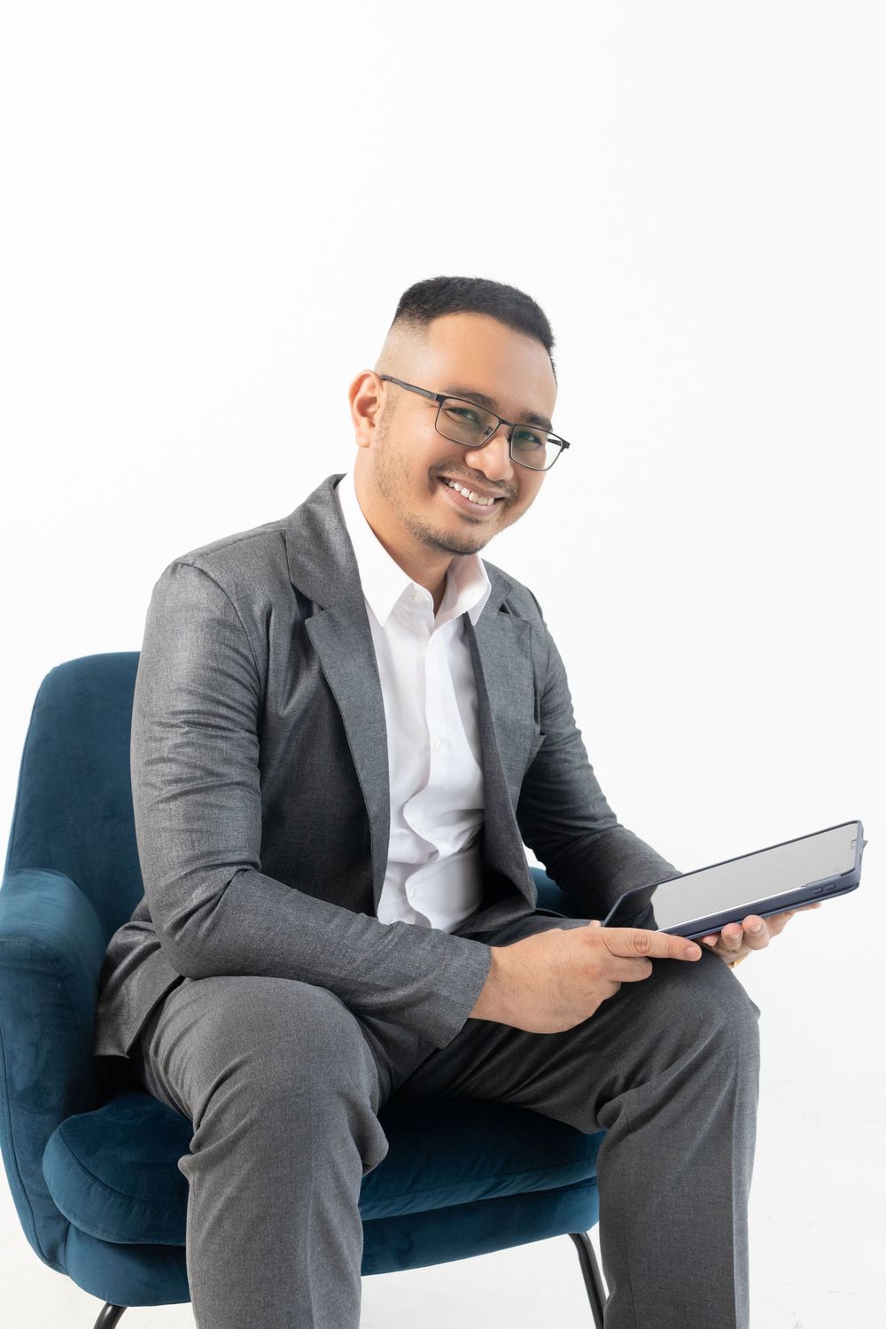 Smiling businessman in gray suit and glasses sitting in blue chair holding a notebook on white background.