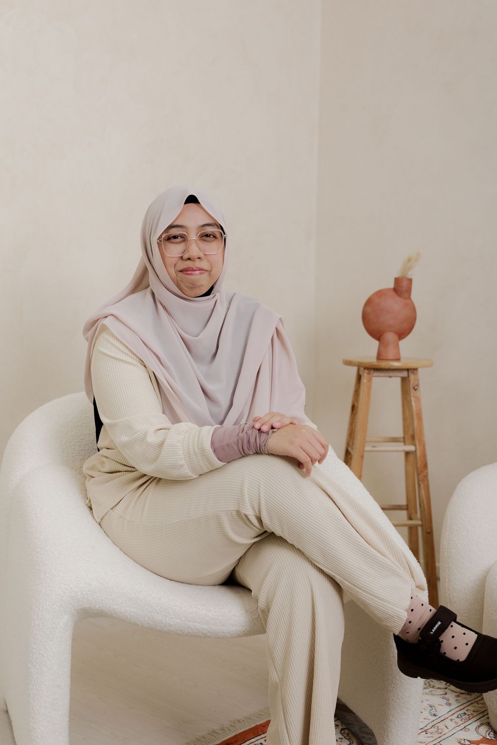 A woman in a light hijab and cream outfit sits gracefully in a white chair beside a wooden stool with a vase.