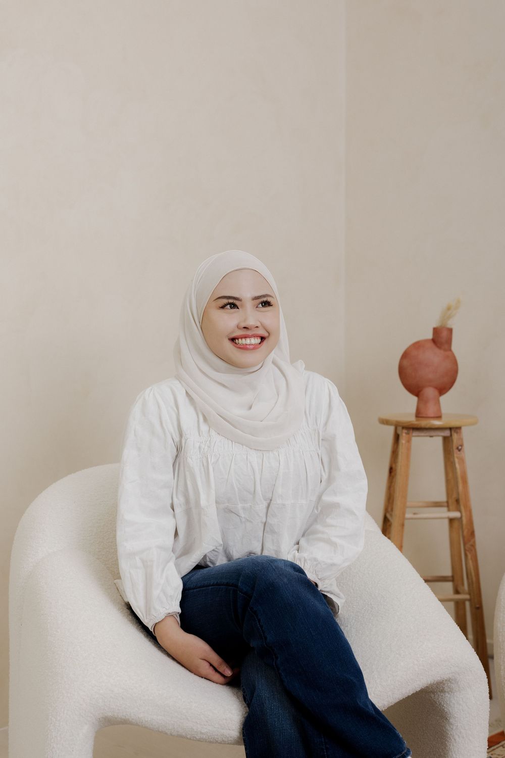 Smiling woman in white hijab and blouse sitting in a modern chair against a neutral beige wall.
