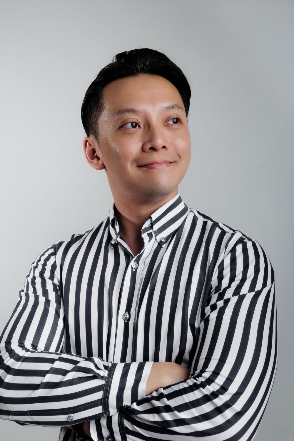 A confident Asian man in a black and white striped shirt stands with arms crossed against a gray background.