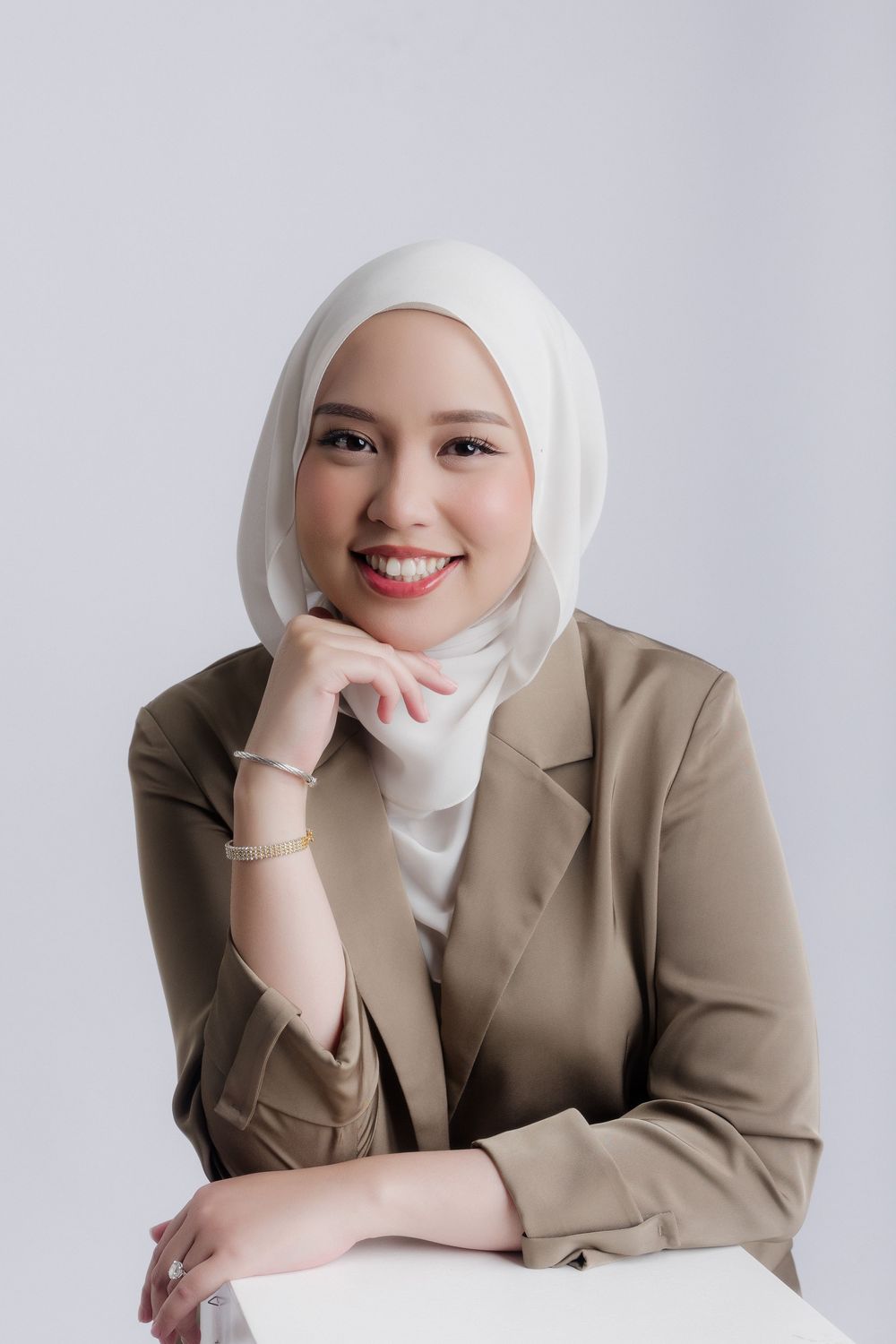 Smiling young Muslim woman in white hijab and tan blazer posing confidently against a light gray background.
