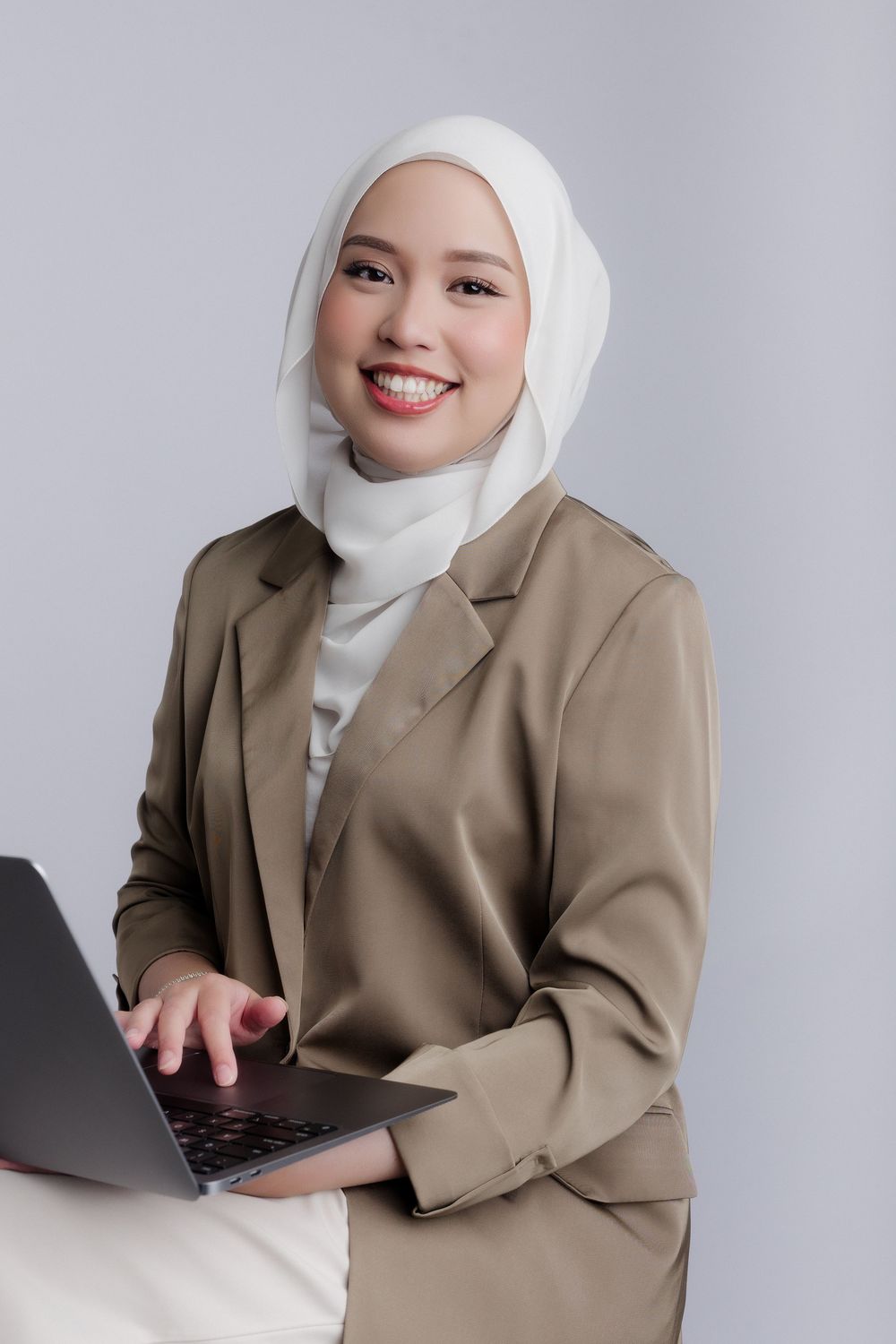 Smiling young Muslim businesswoman in hijab and blazer holding laptop against gray background.