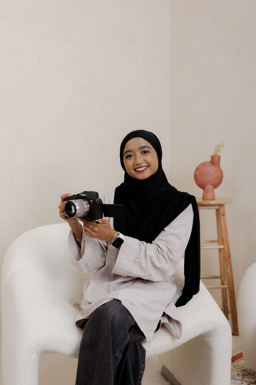 A smiling woman in a black hijab sits in a white chair holding a DSLR camera in a minimalist studio setting.