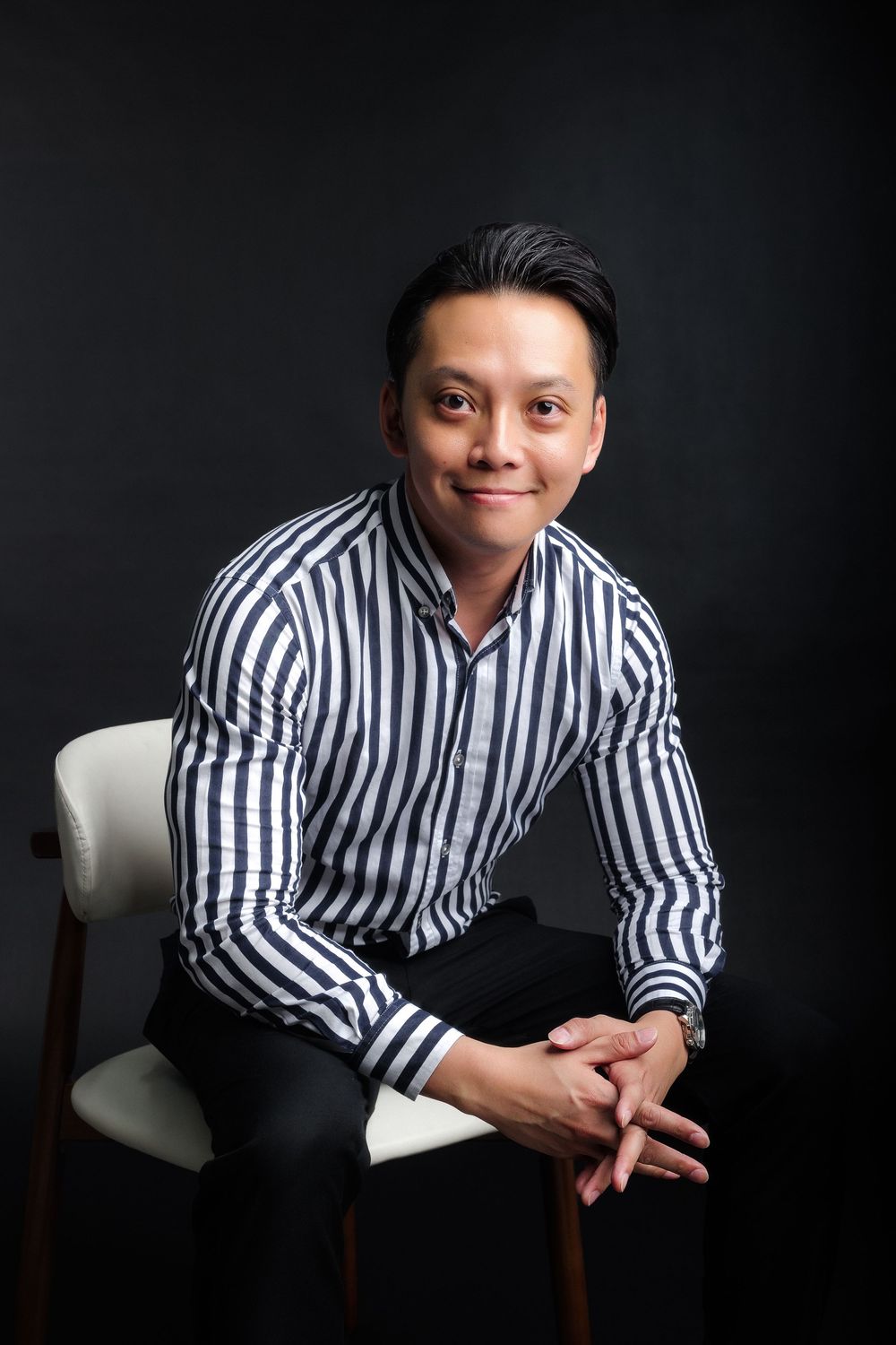 A smiling Asian man in a striped shirt sits relaxed on a white chair against a dark background.