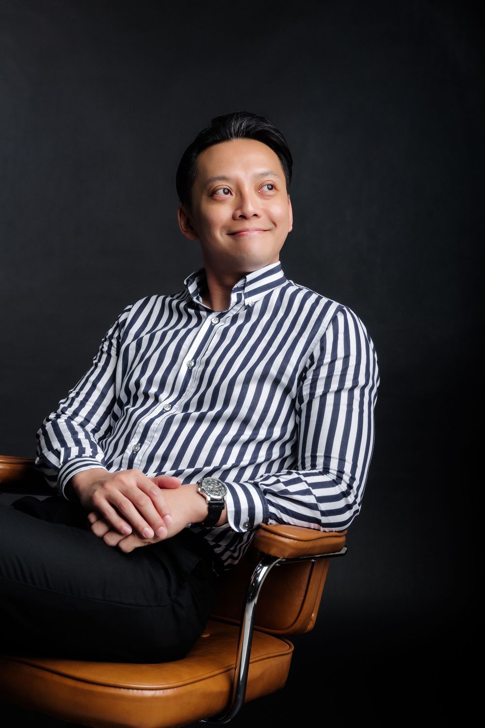 A smiling Asian man in a striped shirt sits relaxed in a leather chair against a dark background.