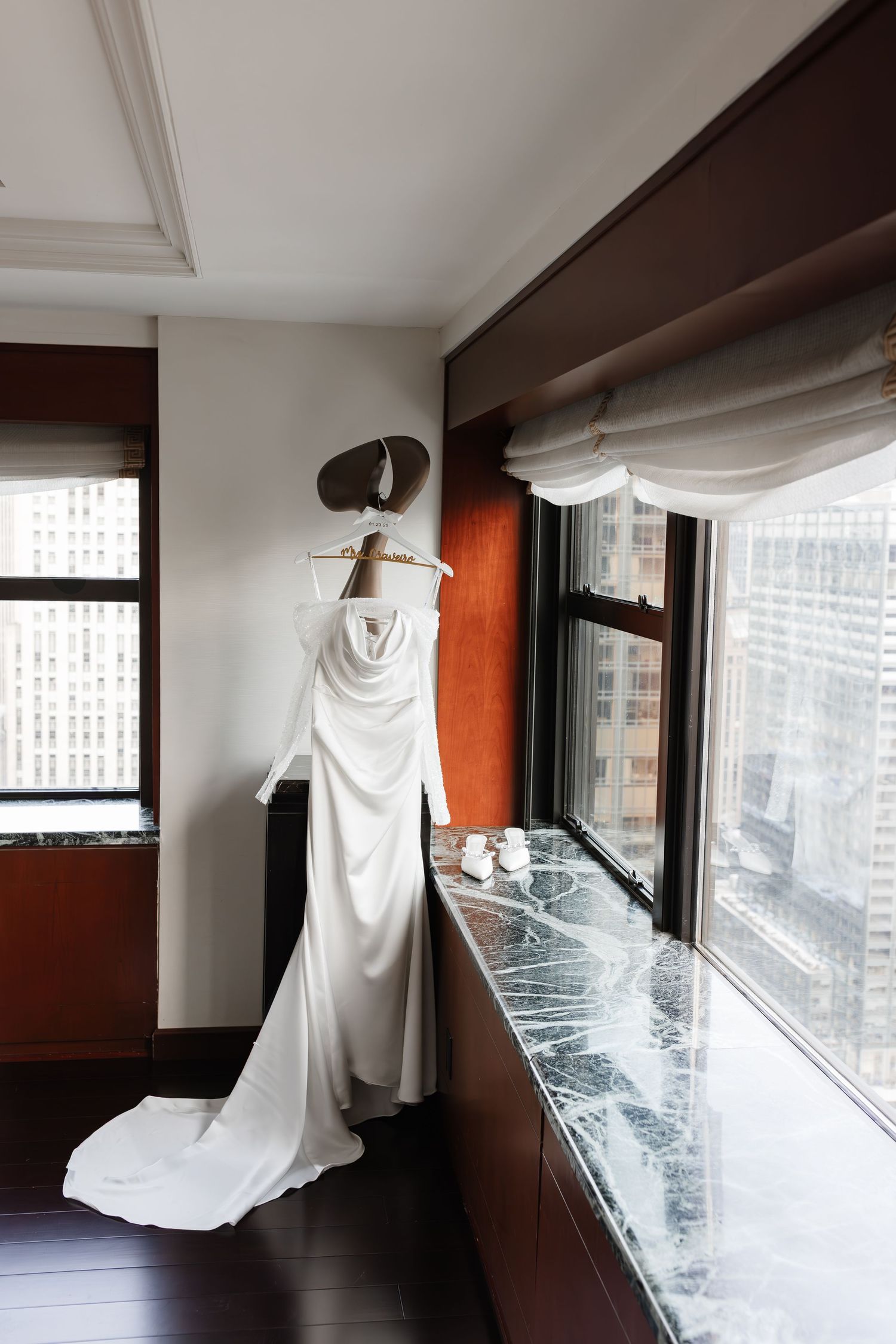 Elegant white bridal gown with long train hanging by a window in a luxury hotel room with city views.