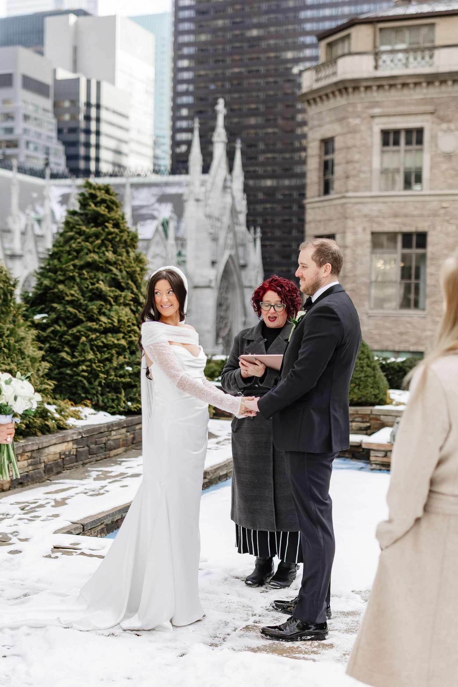 Bride and groom exchange vows outdoors in winter snow with officiant, NYC skyline and stone church backdrop.