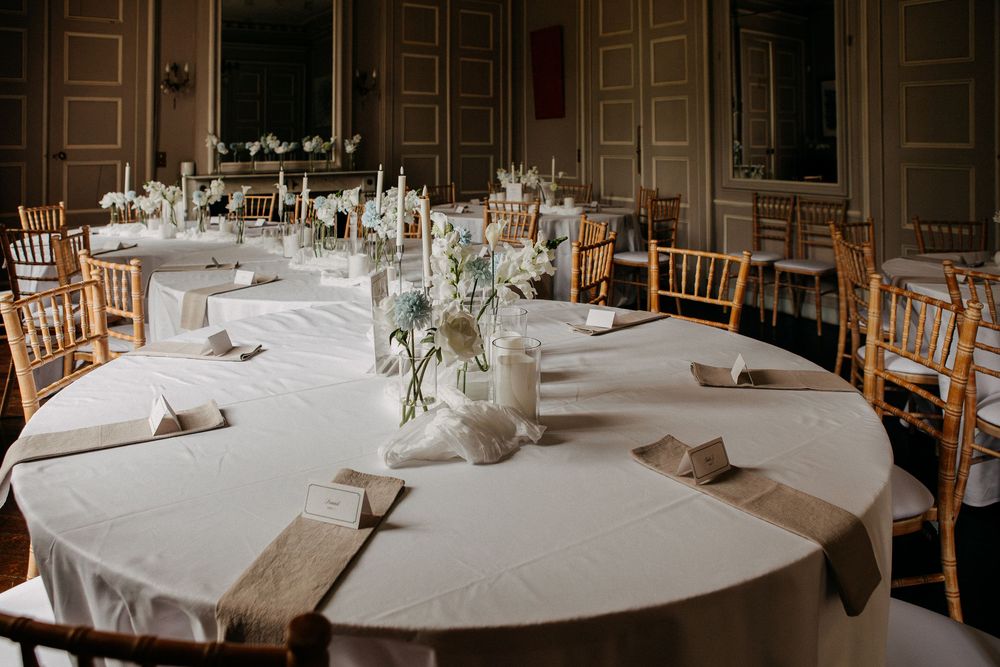 Elegant wedding reception hall with round tables, white linens, floral centerpieces, and gold Chiavari chairs.