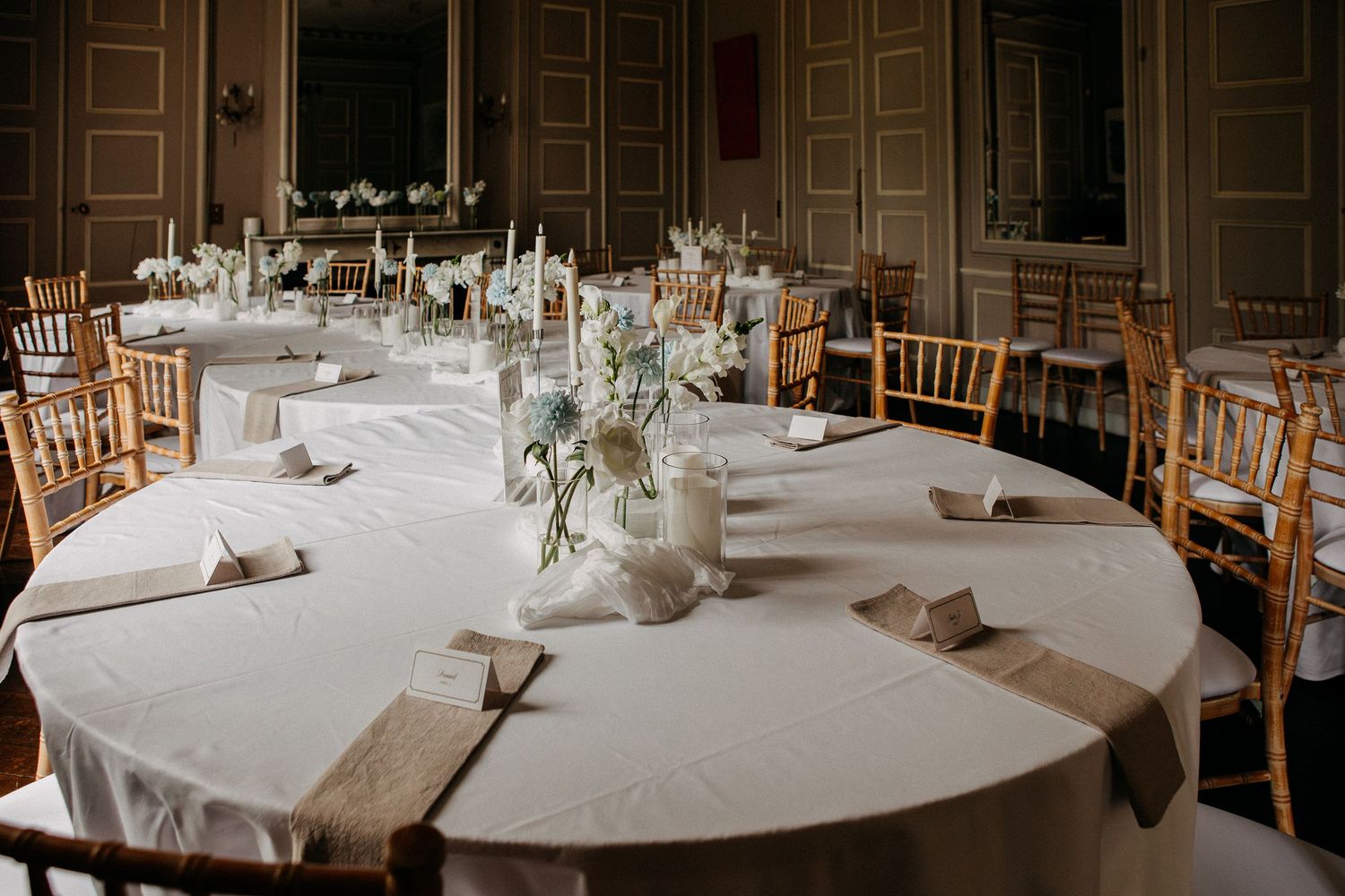 Elegant wedding reception hall with round tables, white linens, floral centerpieces, and gold Chiavari chairs.