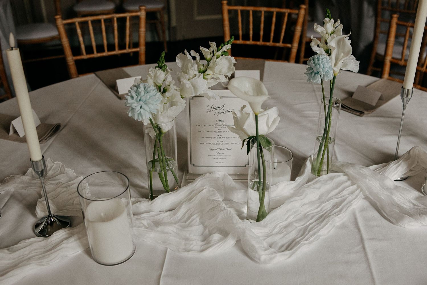 Elegant wedding table centerpiece with white flowers in bud vases, taper candles, and a menu card on white linen.